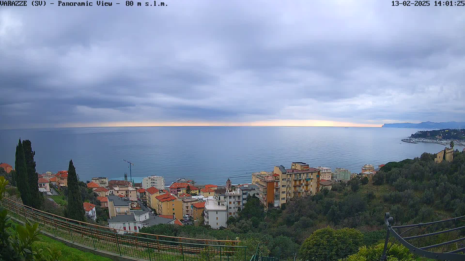 A coastal town with multi-story buildings is nestled on a hillside overlooking a calm sea under a cloudy sky.