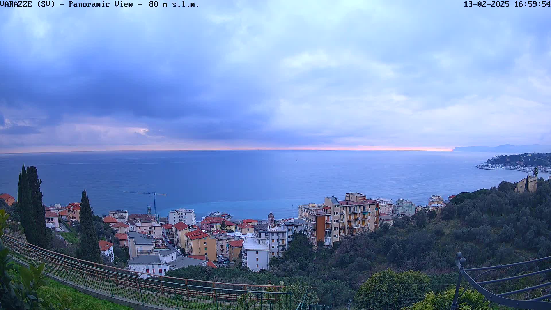 A coastal town with multi-story buildings is nestled on a hillside overlooking a calm sea under a partly cloudy sky.