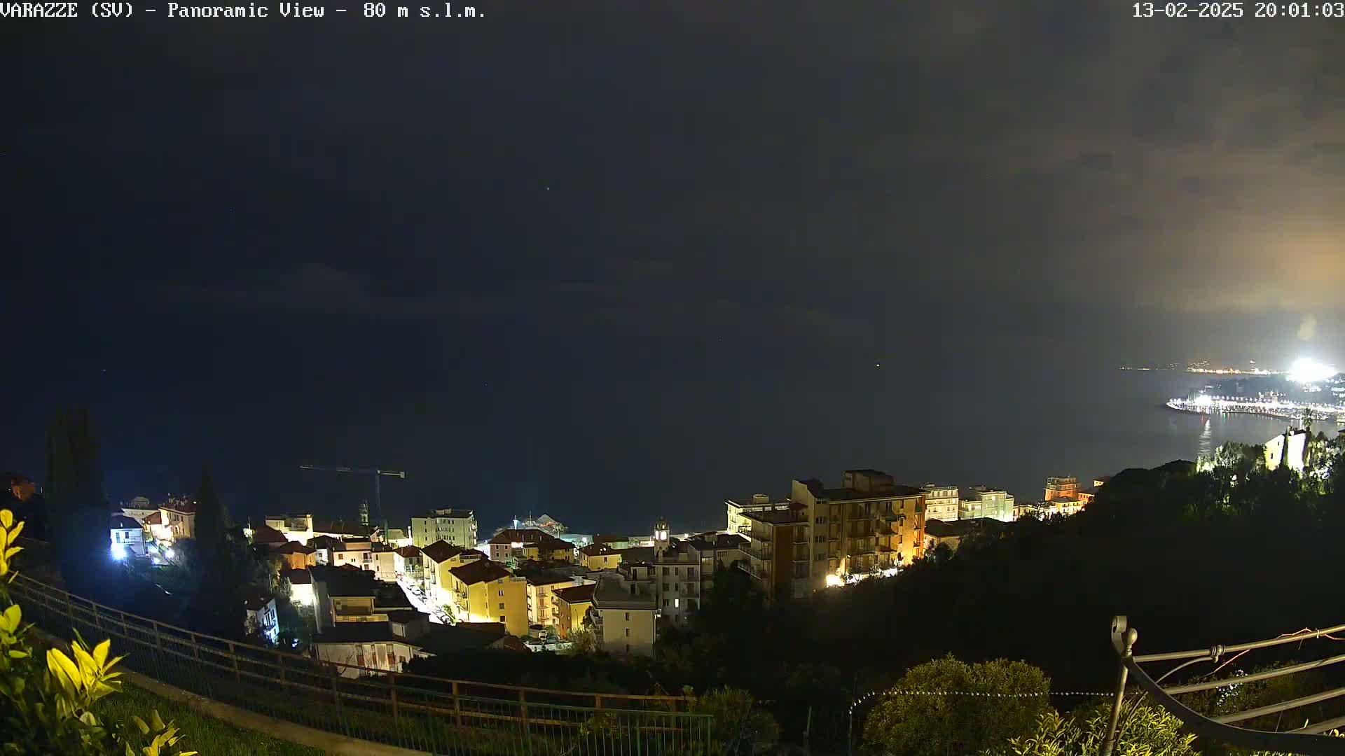 A nighttime panoramic view shows a coastal town illuminated, with a dark, cloudy sky over a calm sea.