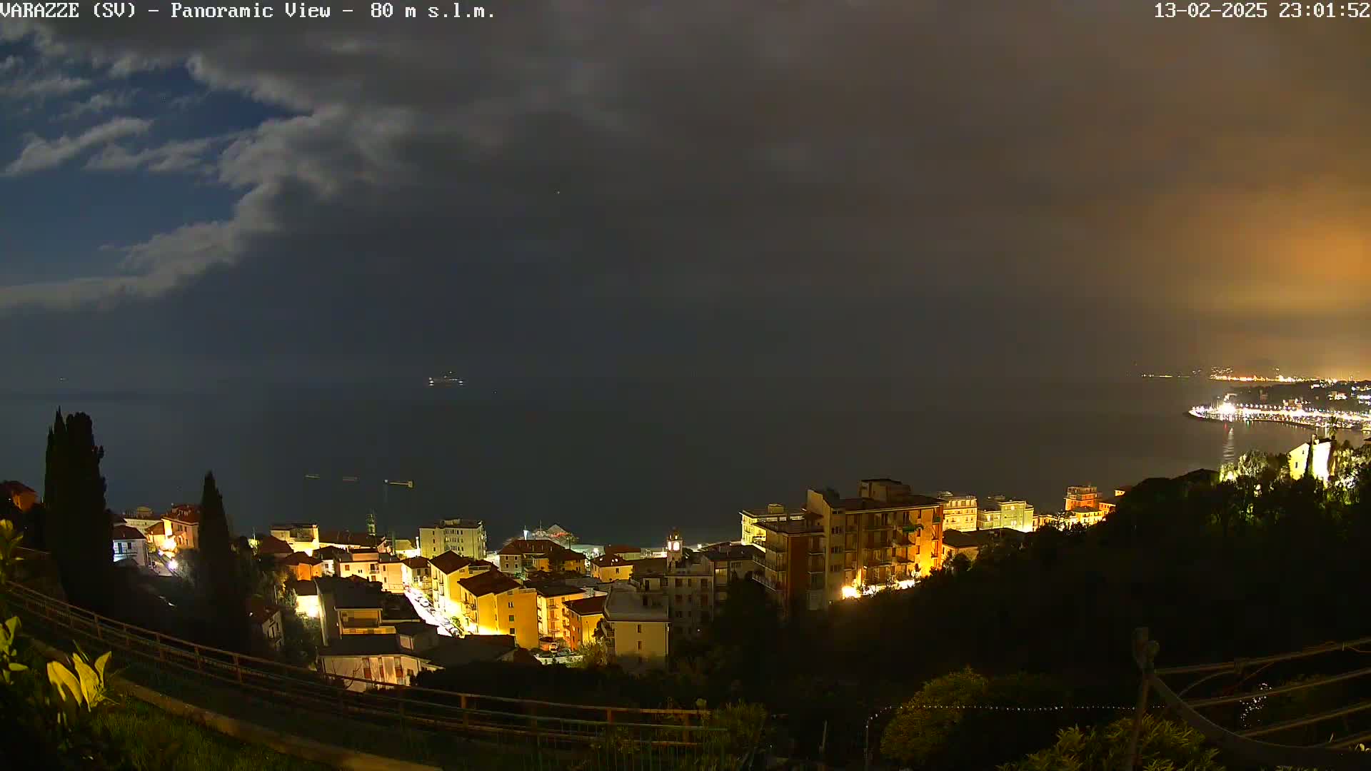 A nighttime panoramic view of a coastal town illuminated with lights, overlooking a dark body of water under a cloudy sky.