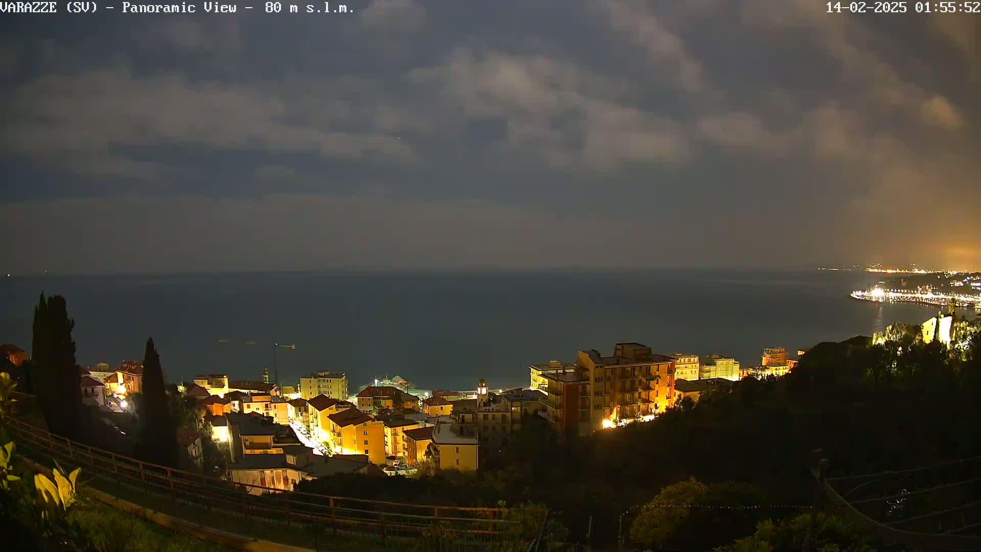 A nighttime panoramic view shows a coastal town with illuminated buildings overlooking a dark sea under a cloudy sky.