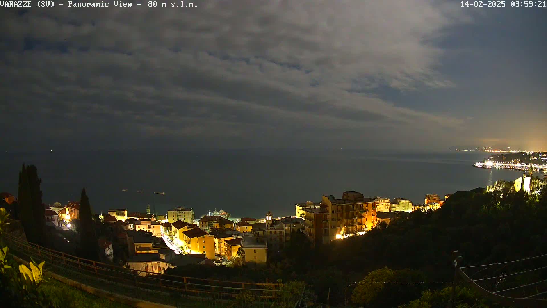 A nighttime panoramic view of a coastal town illuminated with lights, overlooking a calm sea under a partly cloudy sky.