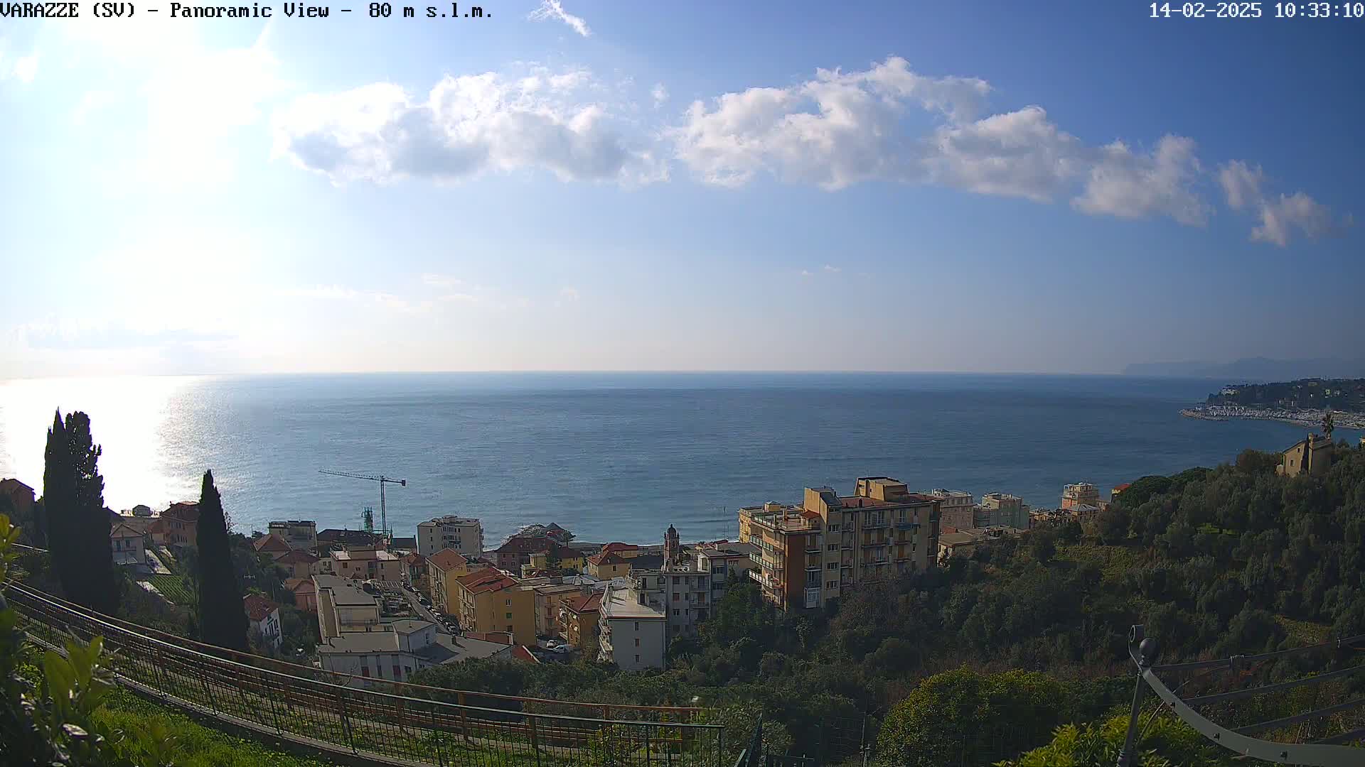 A panoramic view of a coastal town on a partly sunny day, showing buildings, trees, and a calm sea extending to the horizon.
