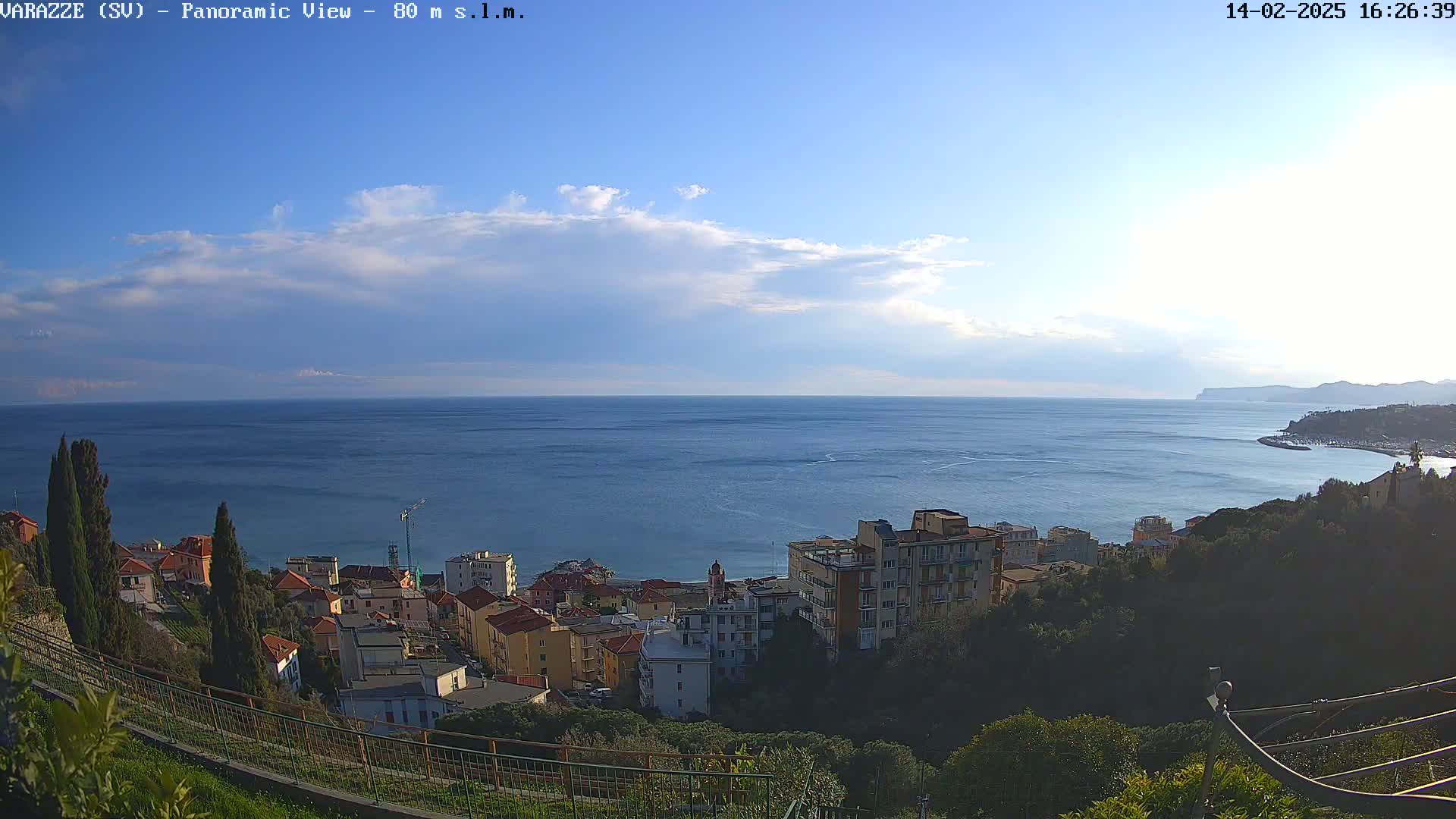 A coastal town nestled on a hillside overlooks a calm, blue sea under a partly cloudy sky.