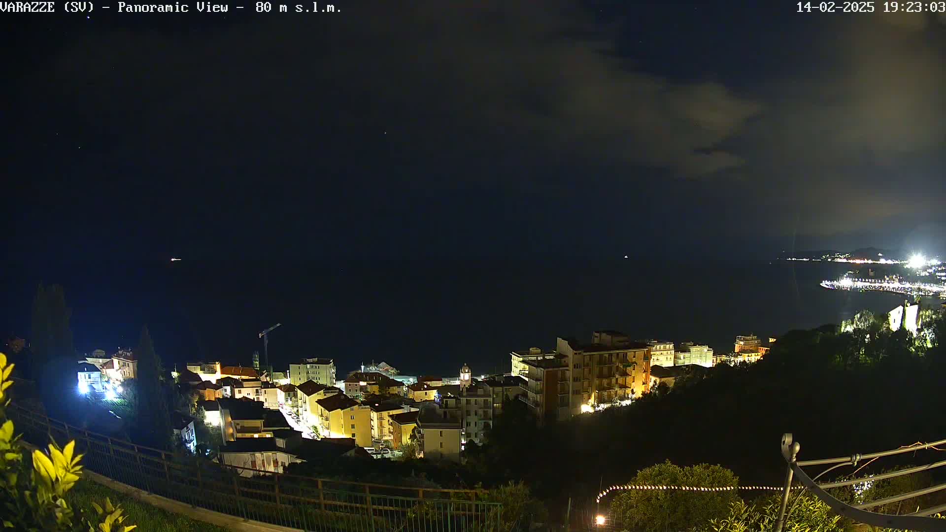 A nighttime, panoramic view shows a coastal town with its lights illuminated against a dark sky with some clouds and a calm sea.