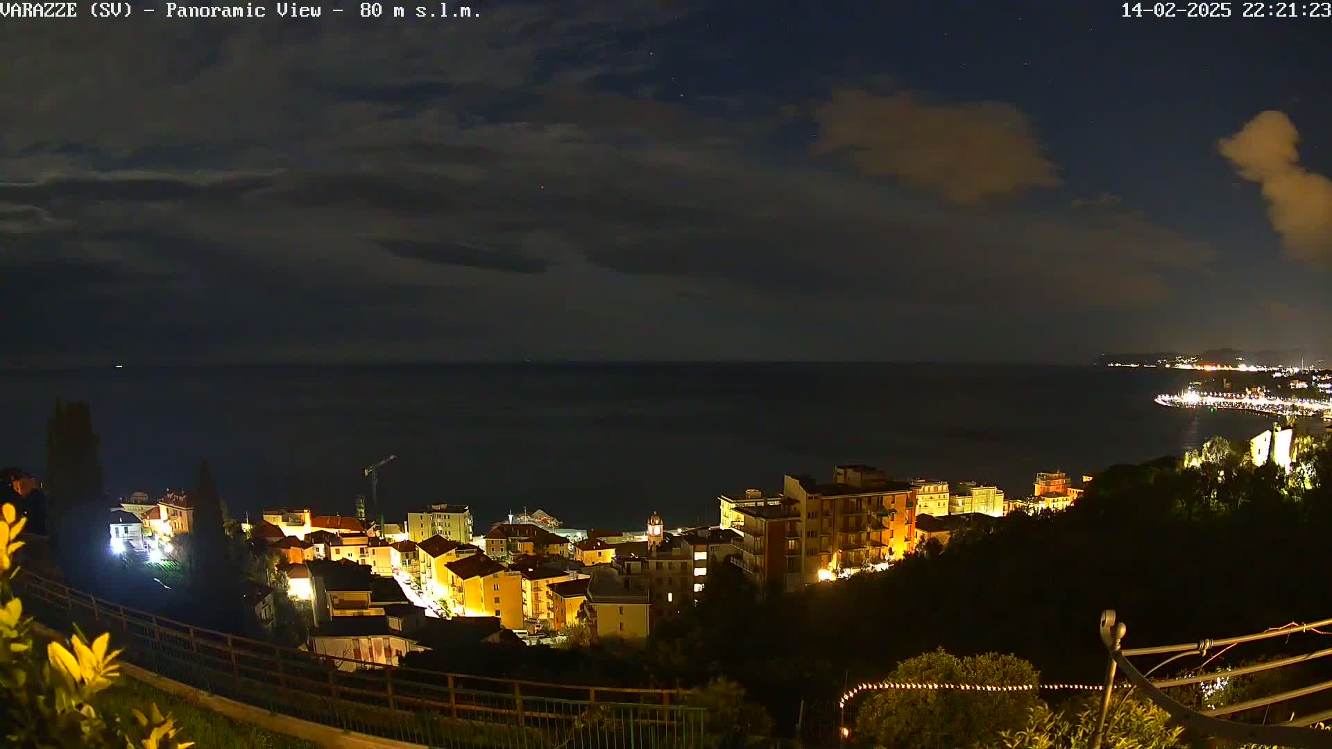A nighttime panoramic view of a coastal town illuminated with lights under a partly cloudy sky.
