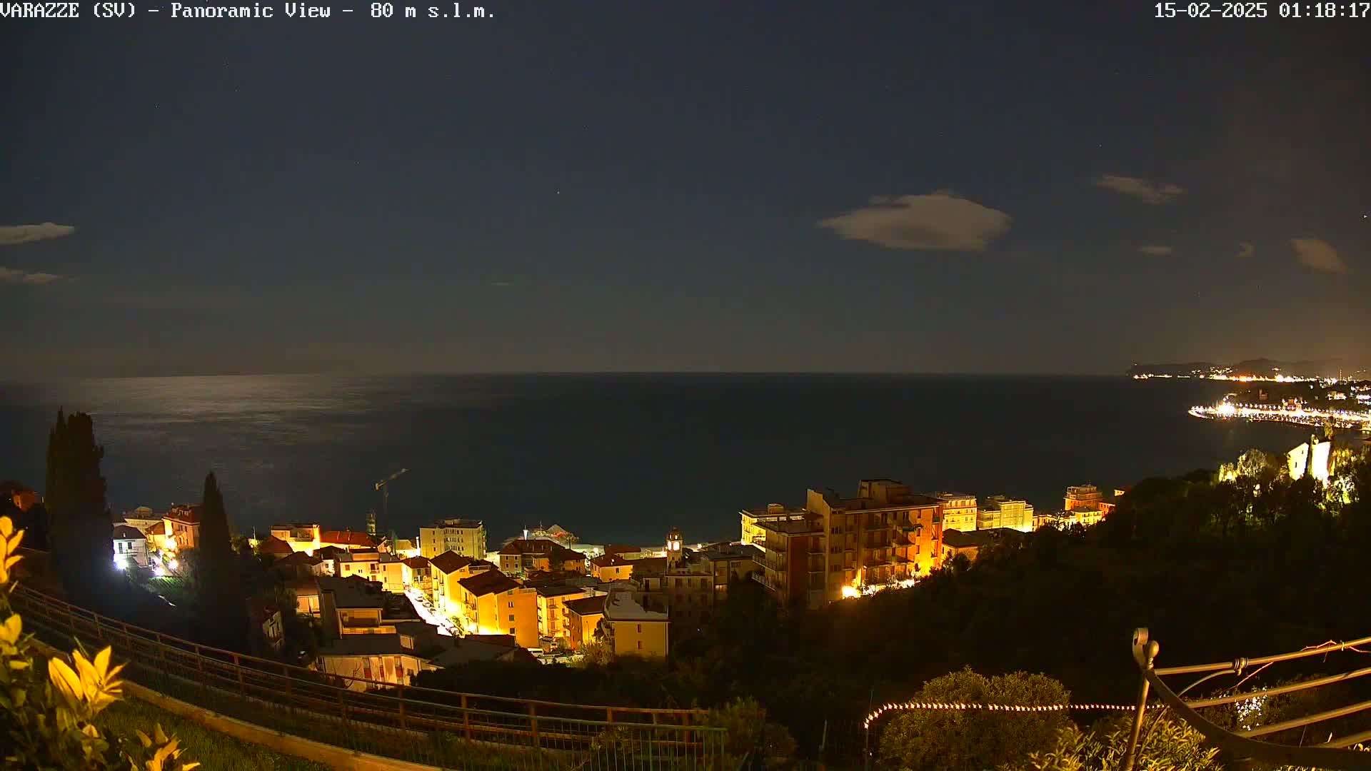 A nighttime panoramic view shows a coastal town illuminated with lights, overlooking a dark sea under a partly cloudy sky.