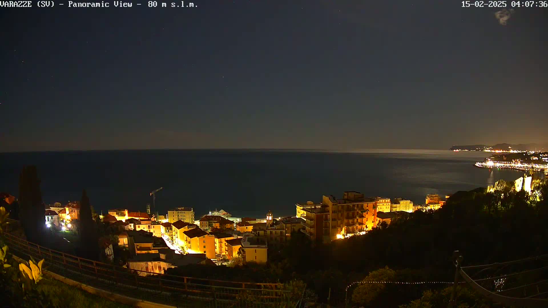 A nighttime panoramic view shows a coastal town with illuminated buildings overlooking a dark sea under a clear, starlit sky.
