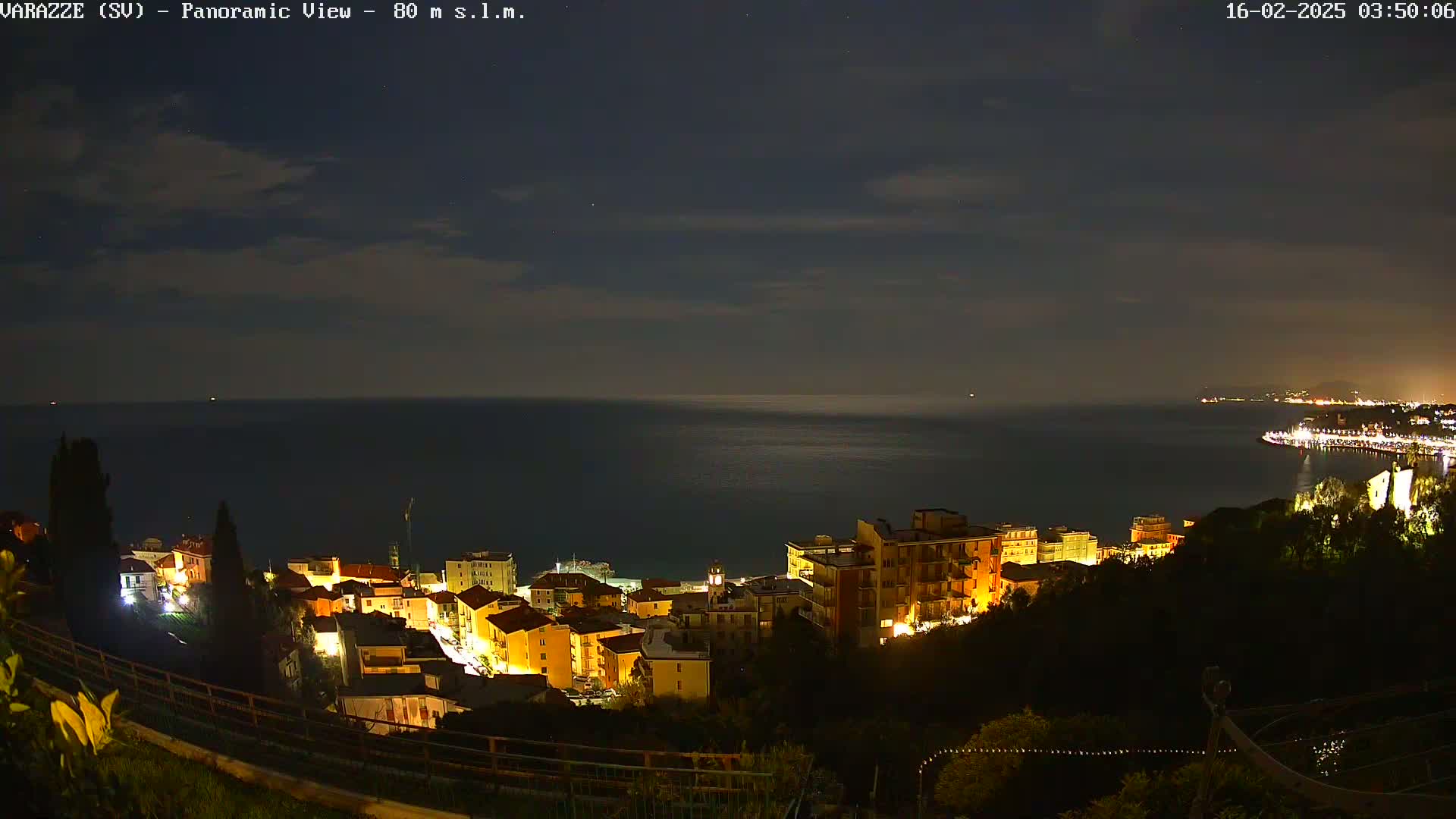 A nighttime panoramic view of a coastal town illuminated by lights, overlooking a dark sea under a mostly clear sky.