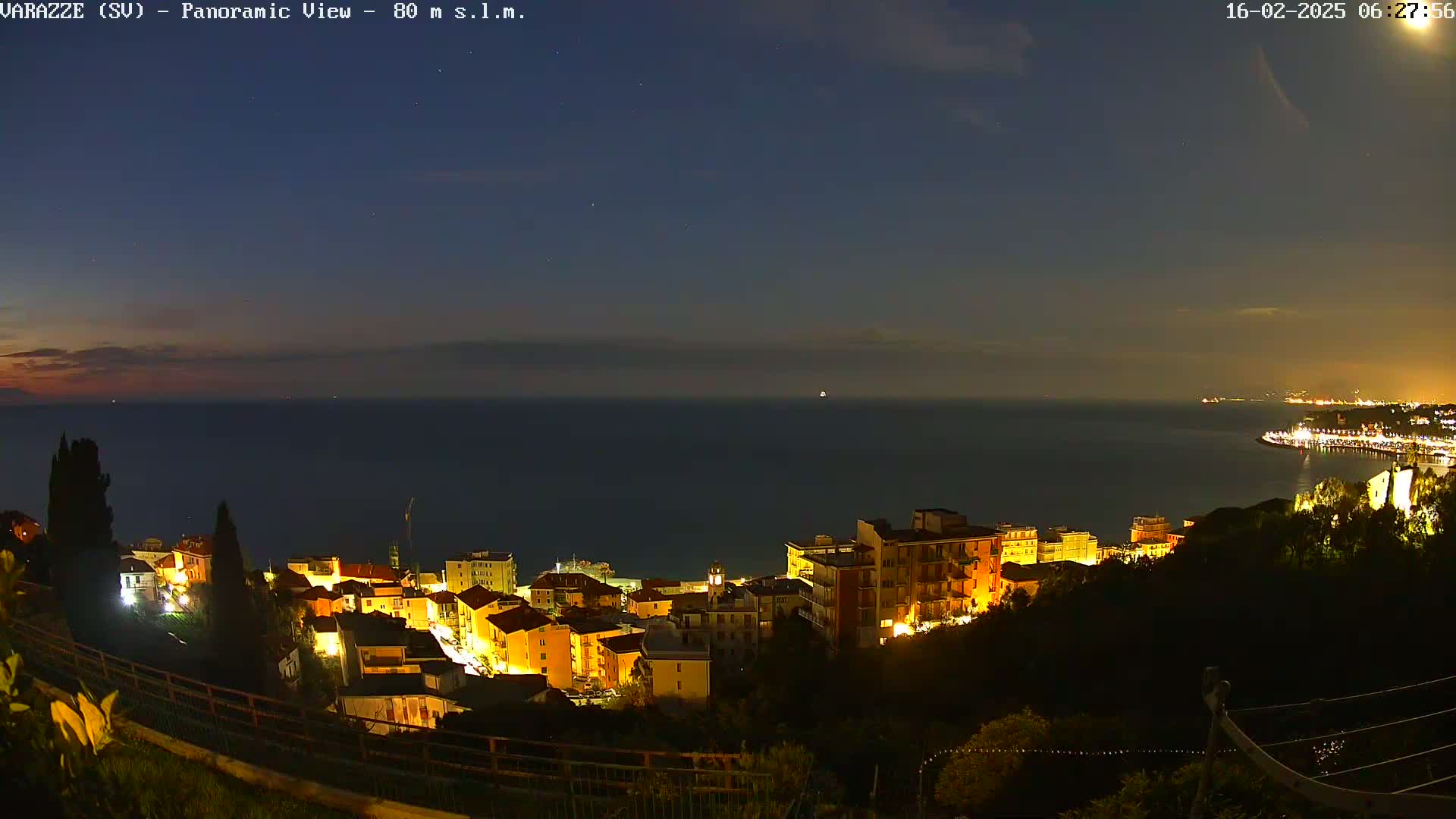 A nighttime panoramic view shows a coastal town illuminated, nestled on a hillside overlooking a dark, calm sea under a mostly clear sky.