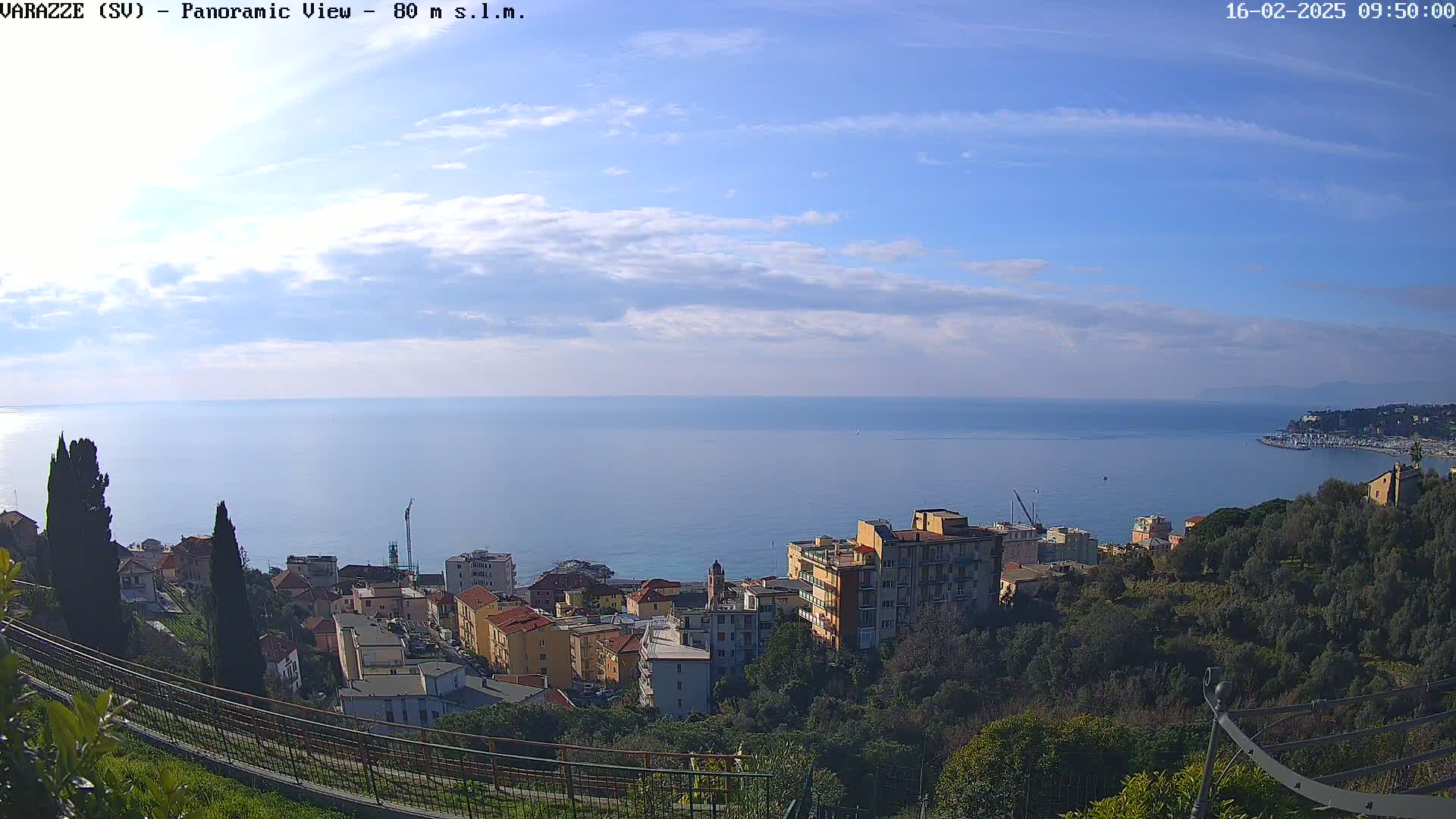A coastal town with multi-story buildings nestled on a hillside overlooking a calm sea under a mostly clear sky with a few scattered clouds.