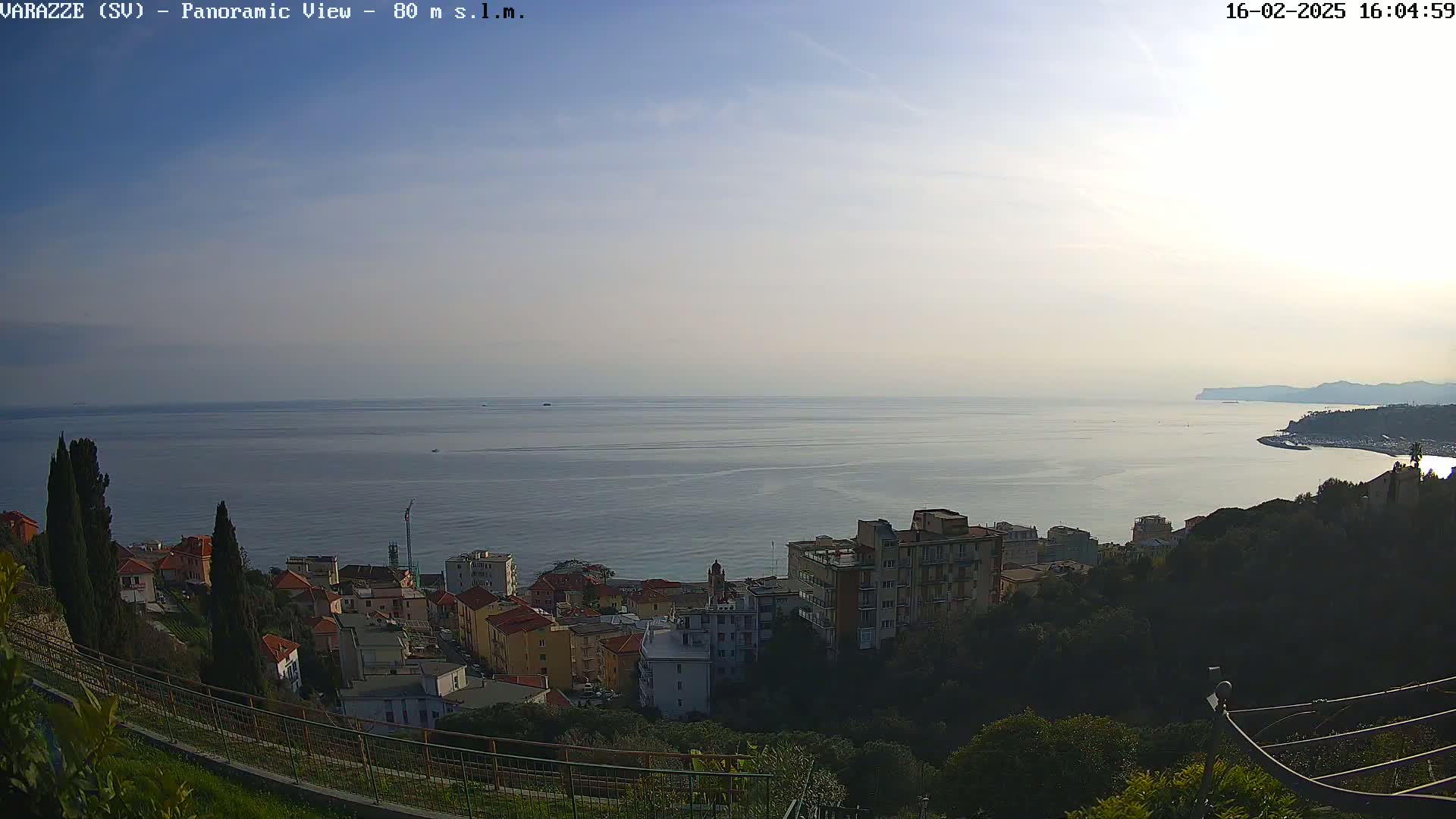 A panoramic view shows a coastal town nestled on a hillside overlooking a calm sea under a mostly clear sky.