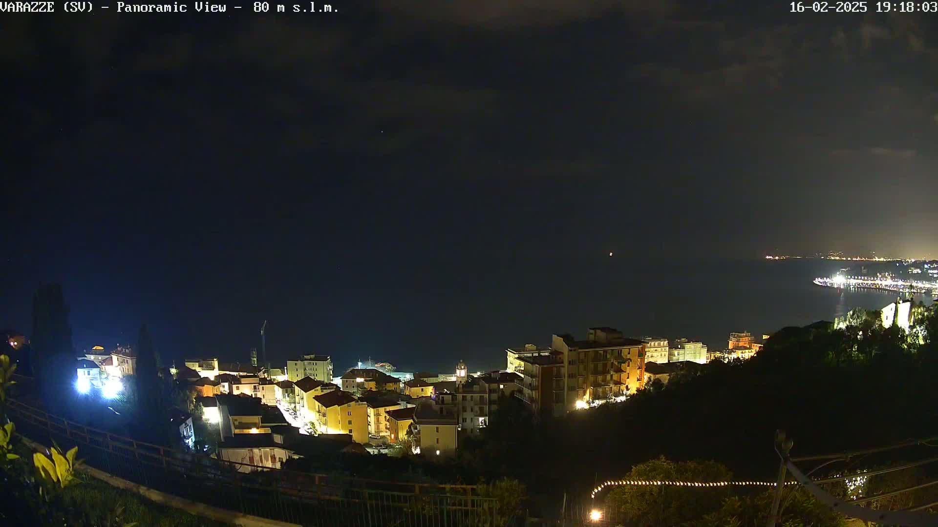 A nighttime panoramic view shows a coastal town illuminated, with a dark, cloudy sky over a calm sea.