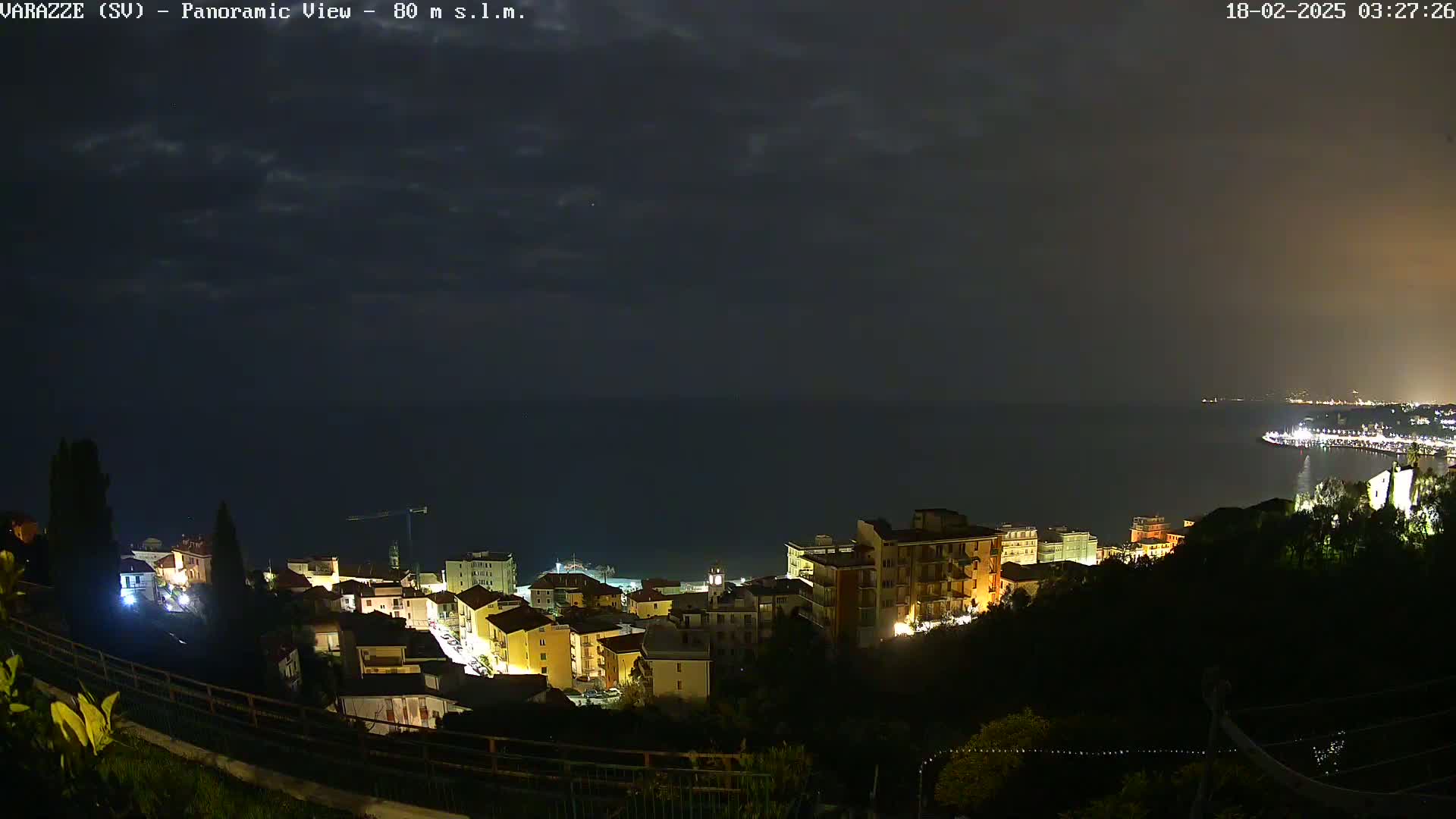 A nighttime panoramic view shows a coastal town with illuminated buildings overlooking a dark ocean under a cloudy sky.