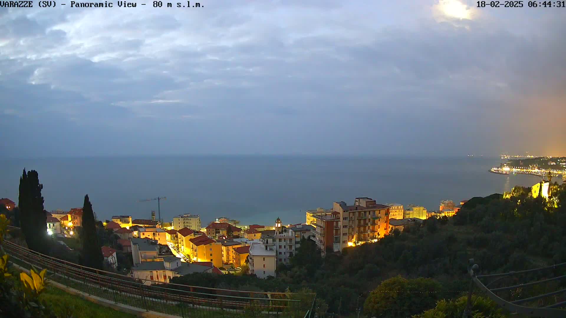 A coastal town at twilight, illuminated with soft lights, overlooks a calm sea under a cloudy sky.
