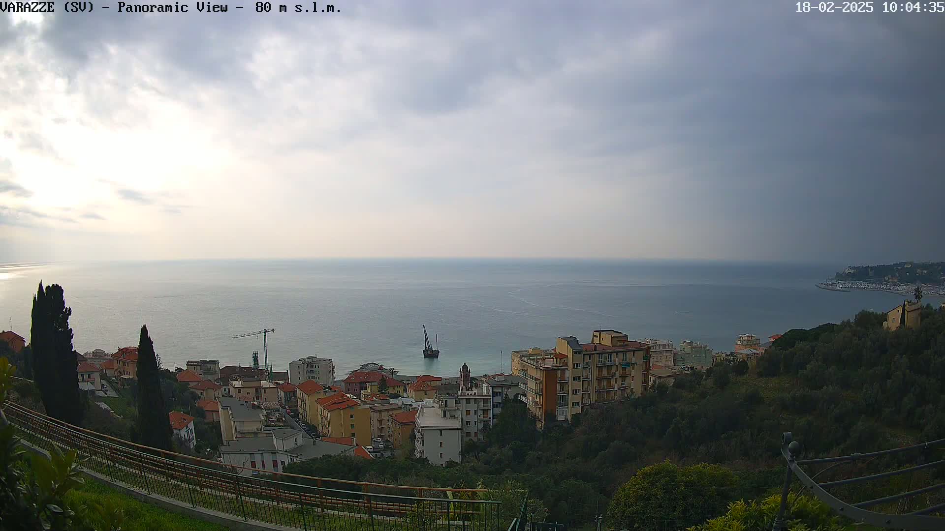 A panoramic view shows a coastal town nestled on a hillside overlooking a calm sea under a cloudy sky.