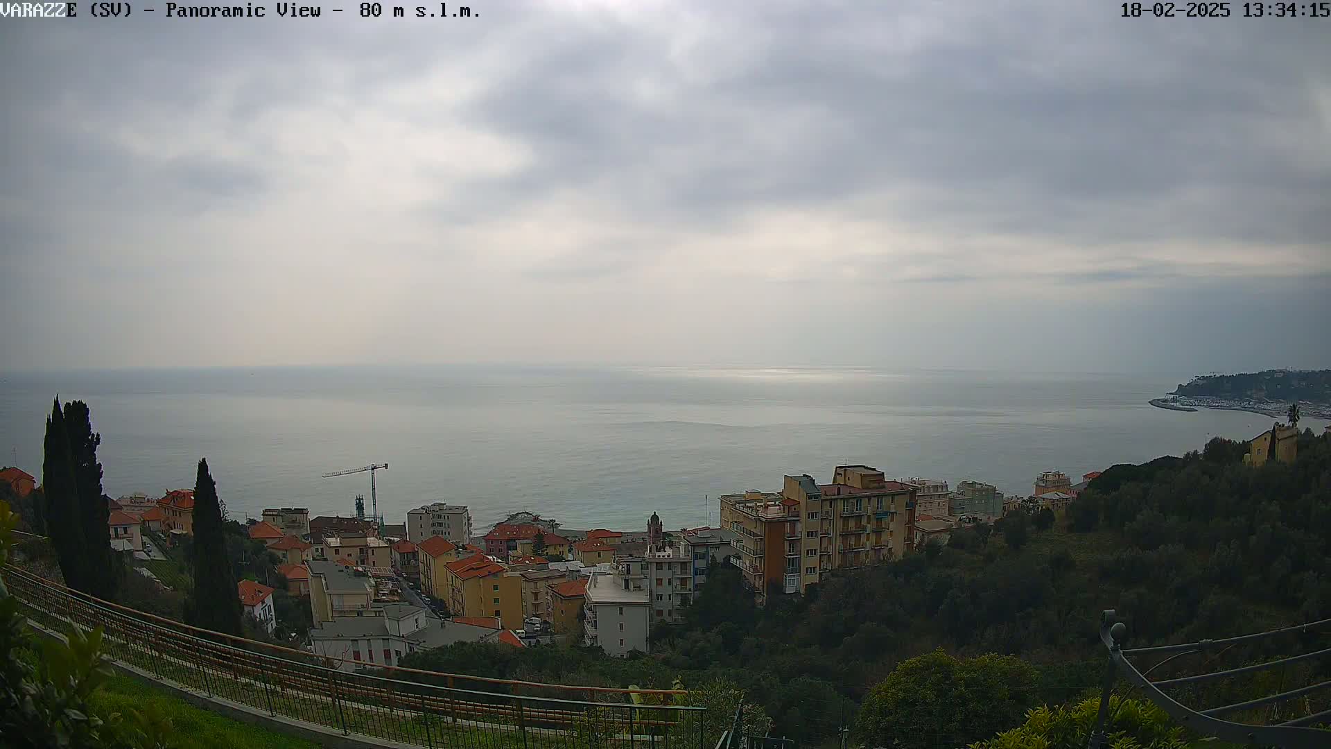 A coastal town with buildings nestled among green hills overlooks a calm sea under a cloudy sky.