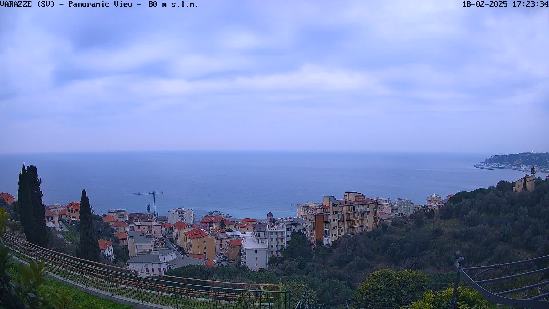 A coastal town nestled on a hillside overlooks a calm sea under a mostly cloudy sky.