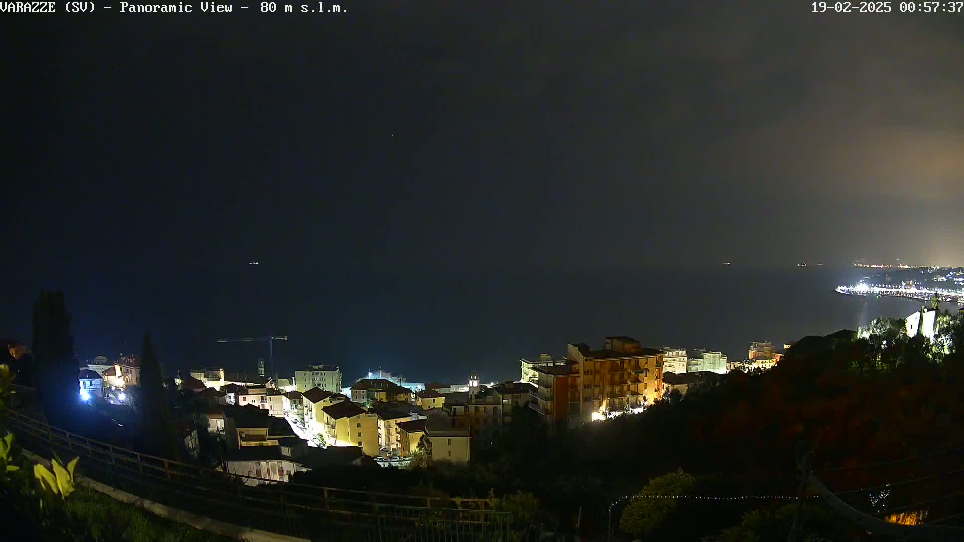 A nighttime panoramic view of a coastal town, illuminated with lights, overlooking a dark sea under a mostly clear sky.