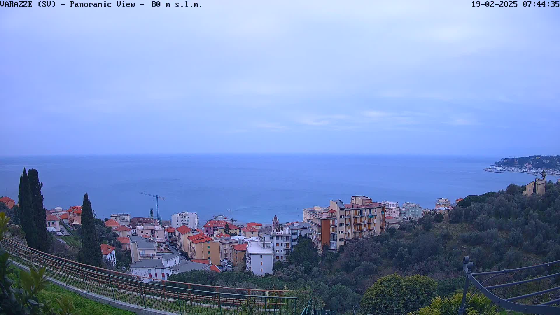 A panoramic view of a coastal town under a cloudy sky, showing buildings and vegetation overlooking a calm sea.