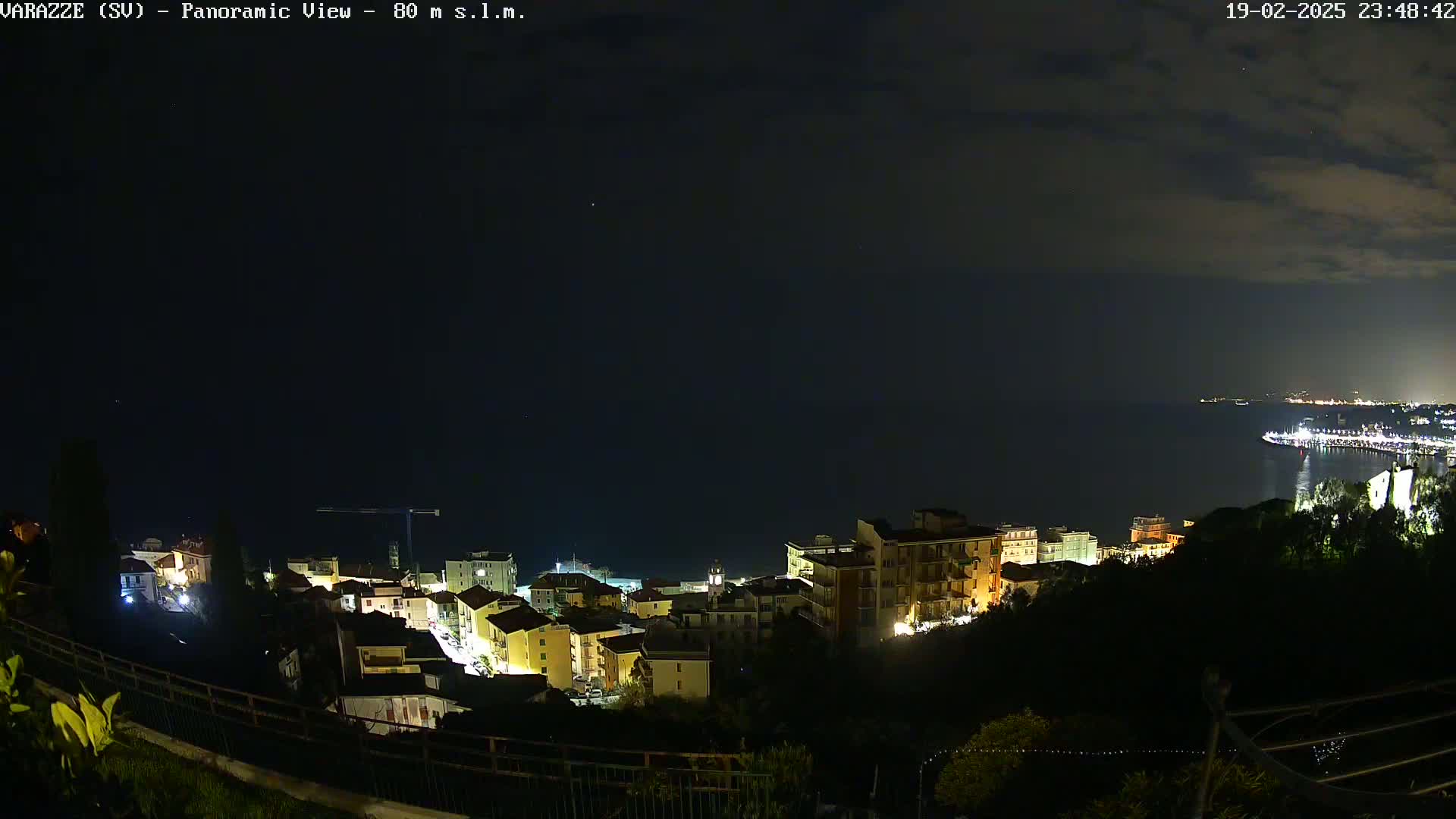 A nighttime panoramic view of a coastal town illuminated by lights, under a partly cloudy sky, overlooking a dark sea.