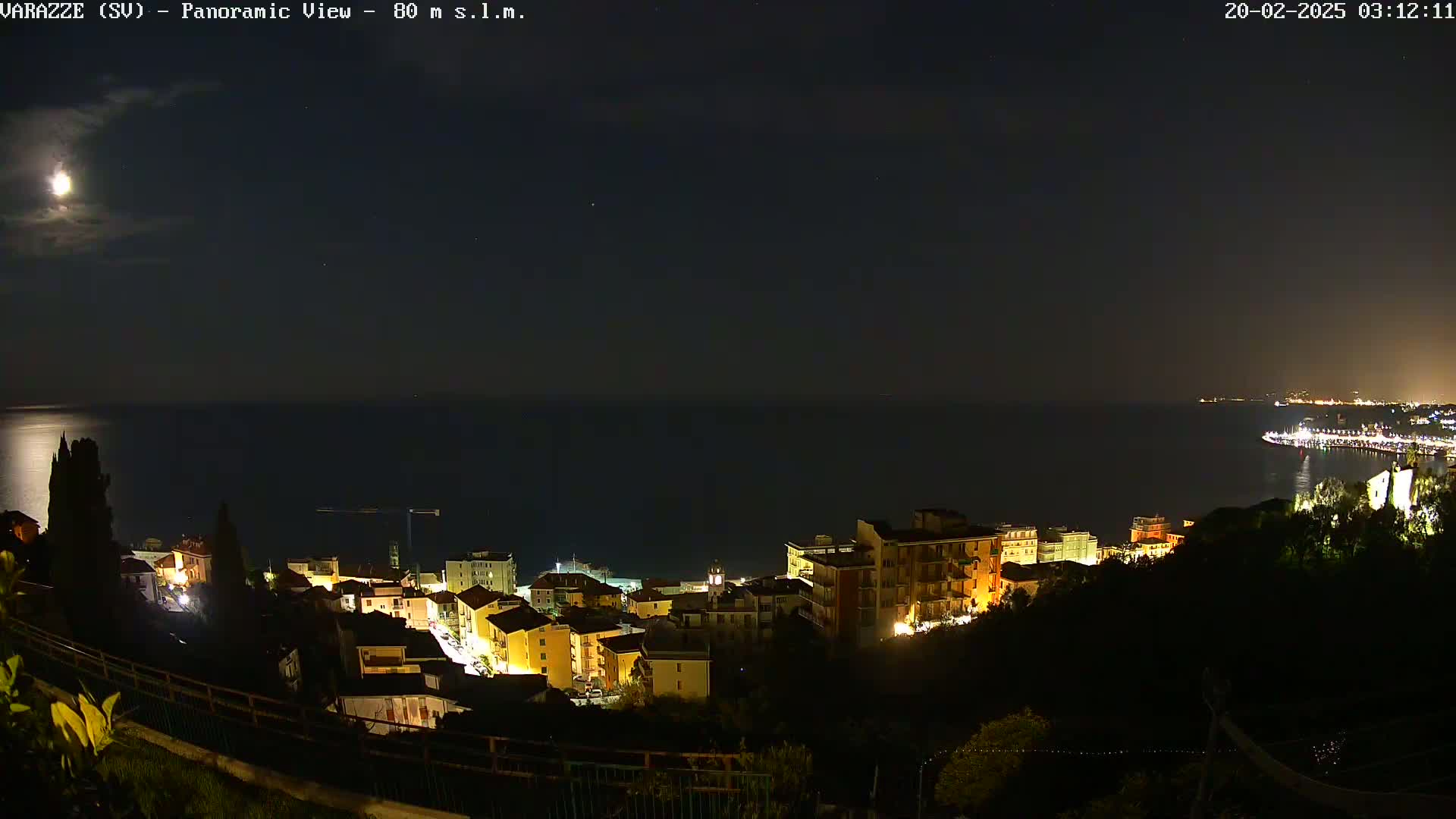 A nighttime panoramic view shows a coastal town illuminated with lights, overlooking a dark sea under a bright moon.