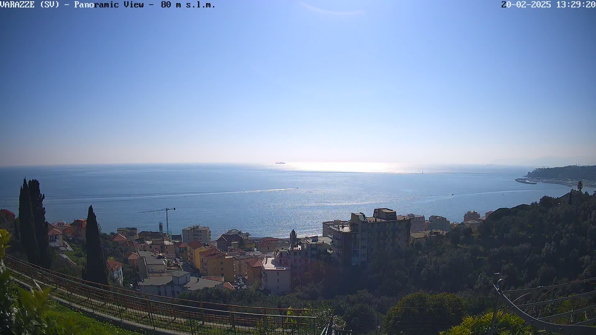 A panoramic view of a coastal town on a sunny day shows a calm sea extending to the horizon, with buildings situated on a hillside overlooking the water.