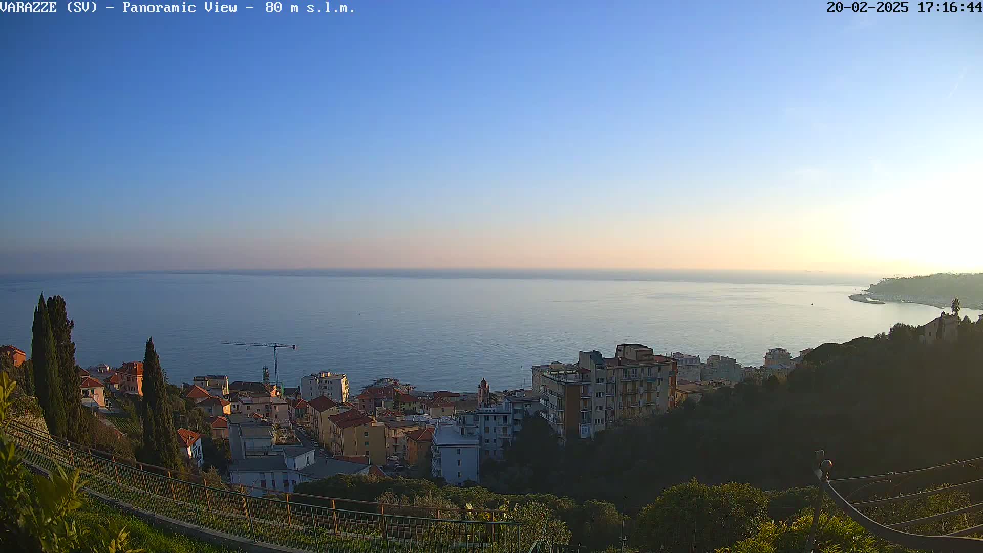 A coastal town with buildings nestled on a hillside overlooks a calm sea under a clear, twilight sky.