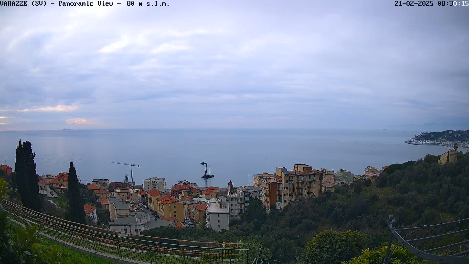 A panoramic view of a coastal town on a cloudy day shows a calm sea, buildings nestled on a hillside, and a distant coastline.