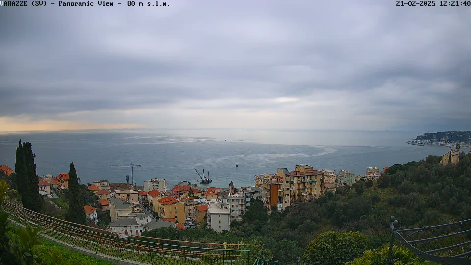 A coastal town with multi-story buildings nestled on a hillside overlooking a calm sea under a cloudy sky.