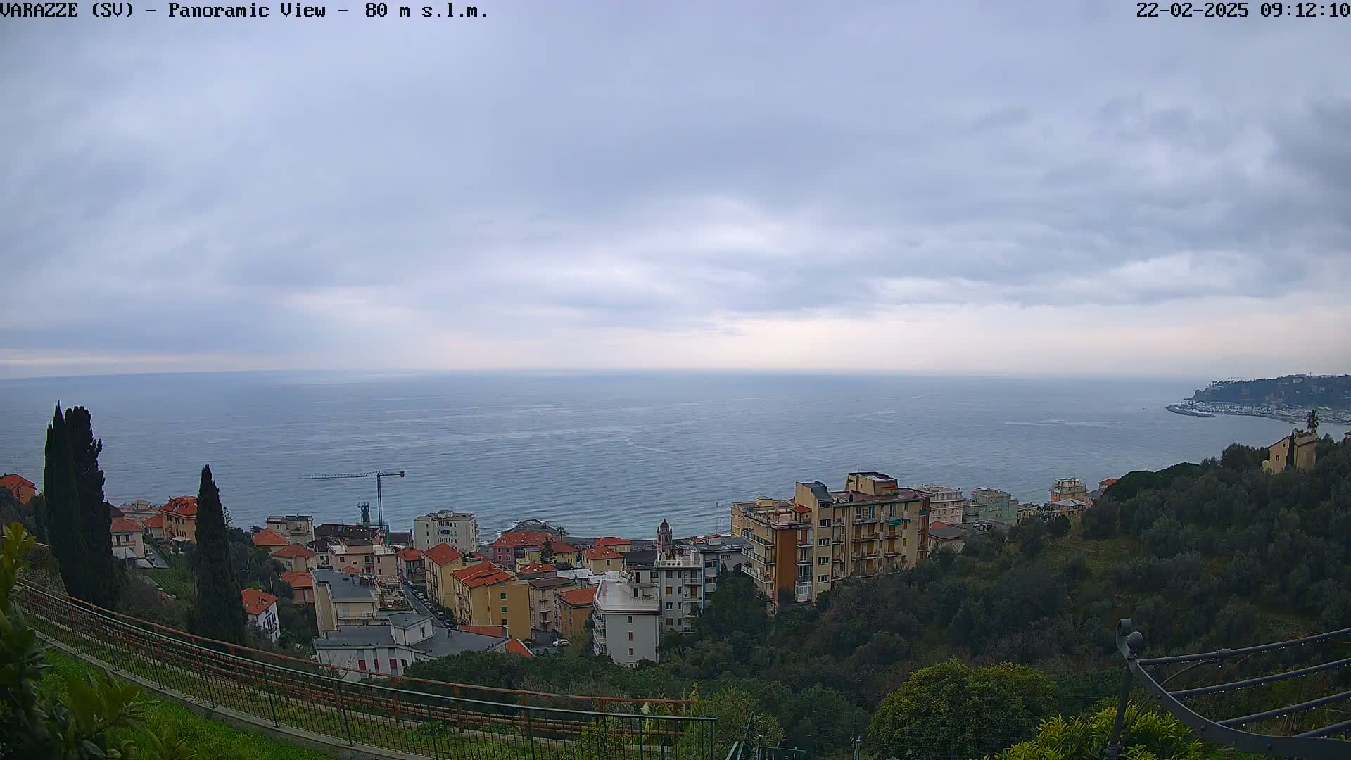 A coastal town with low buildings is nestled on a hillside overlooking a calm sea under an overcast sky.