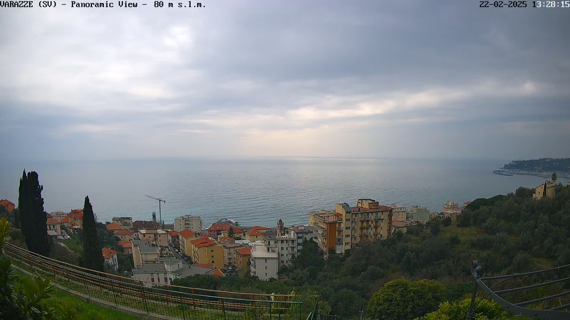A coastal town with multi-story buildings sits on a hillside overlooking a calm sea under an overcast sky.