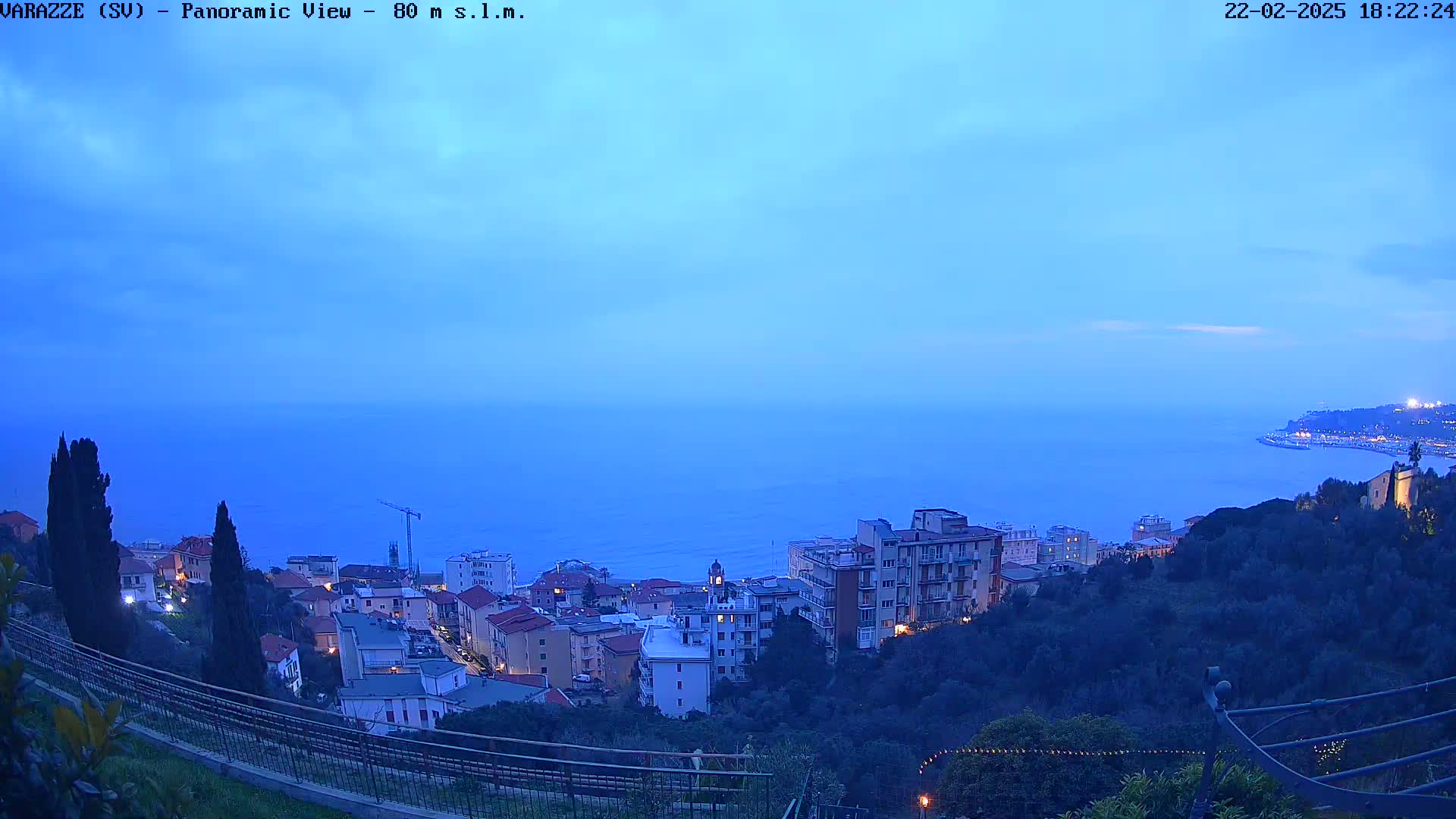 A twilight view overlooking a coastal town nestled on a hillside, with a calm sea extending to the horizon under a clear, dusky sky.