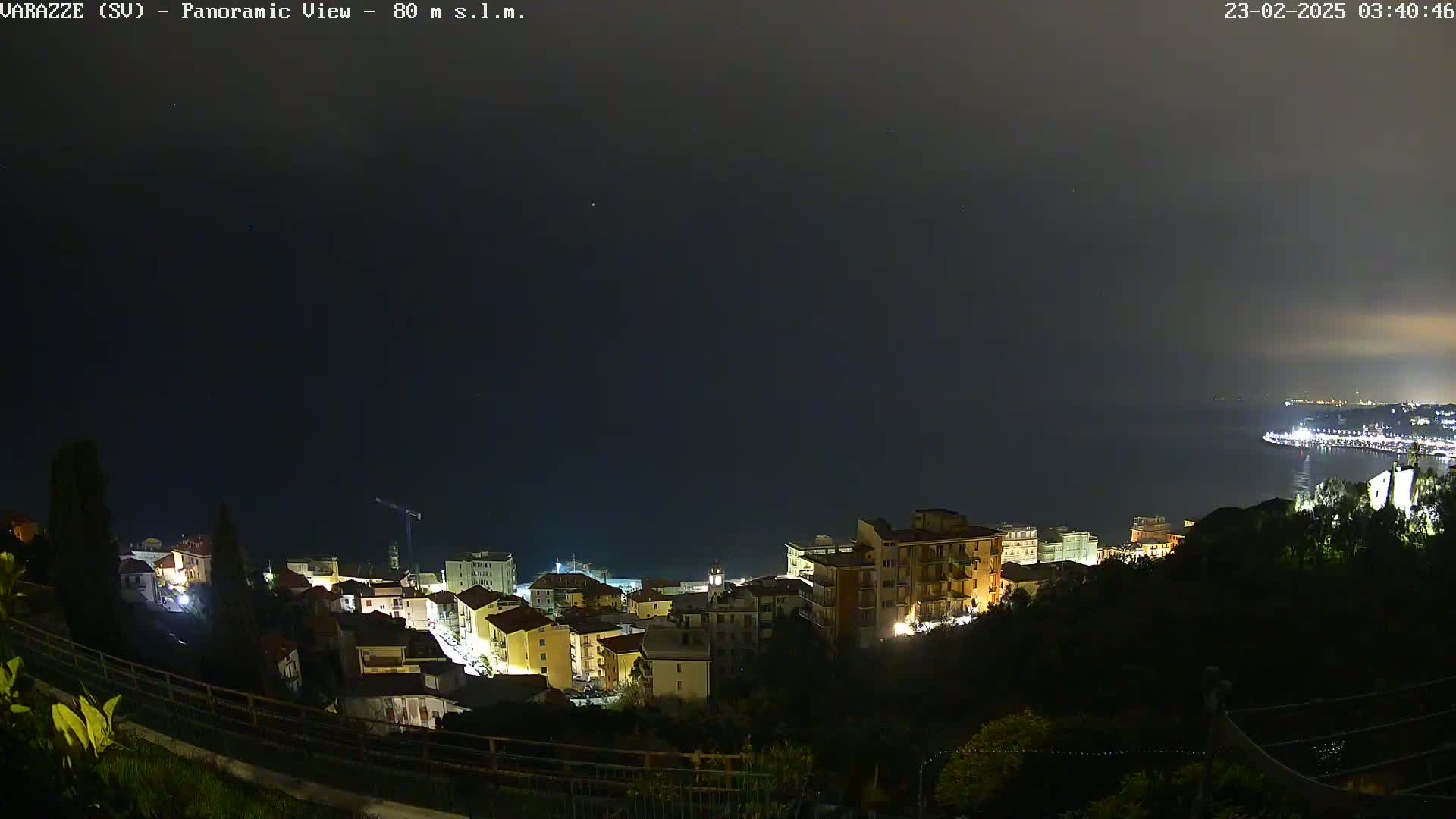 A nighttime panoramic view shows a coastal town illuminated, with a dark, calm sea extending to a dimly lit city in the distance under a cloudy sky.