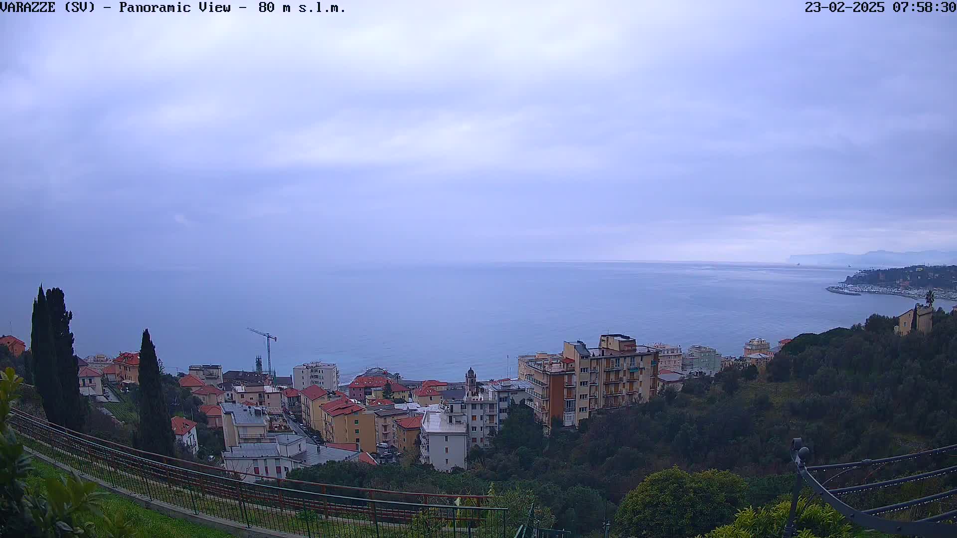 A panoramic view of a coastal town on a cloudy day, showing multi-story buildings, green vegetation, and a calm sea extending to a distant shoreline.