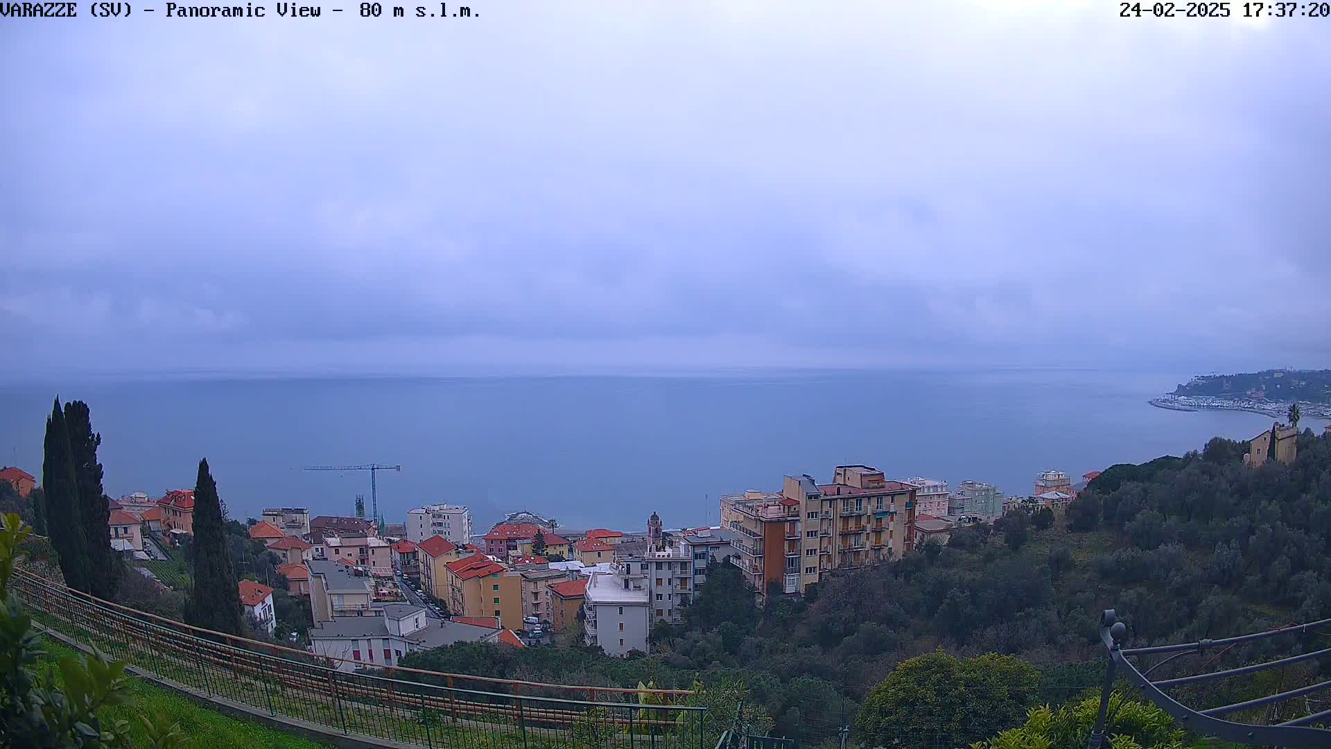 A panoramic view of a coastal town on a cloudy day shows a calm sea extending to the horizon, with buildings and trees cascading down a hillside toward the water.