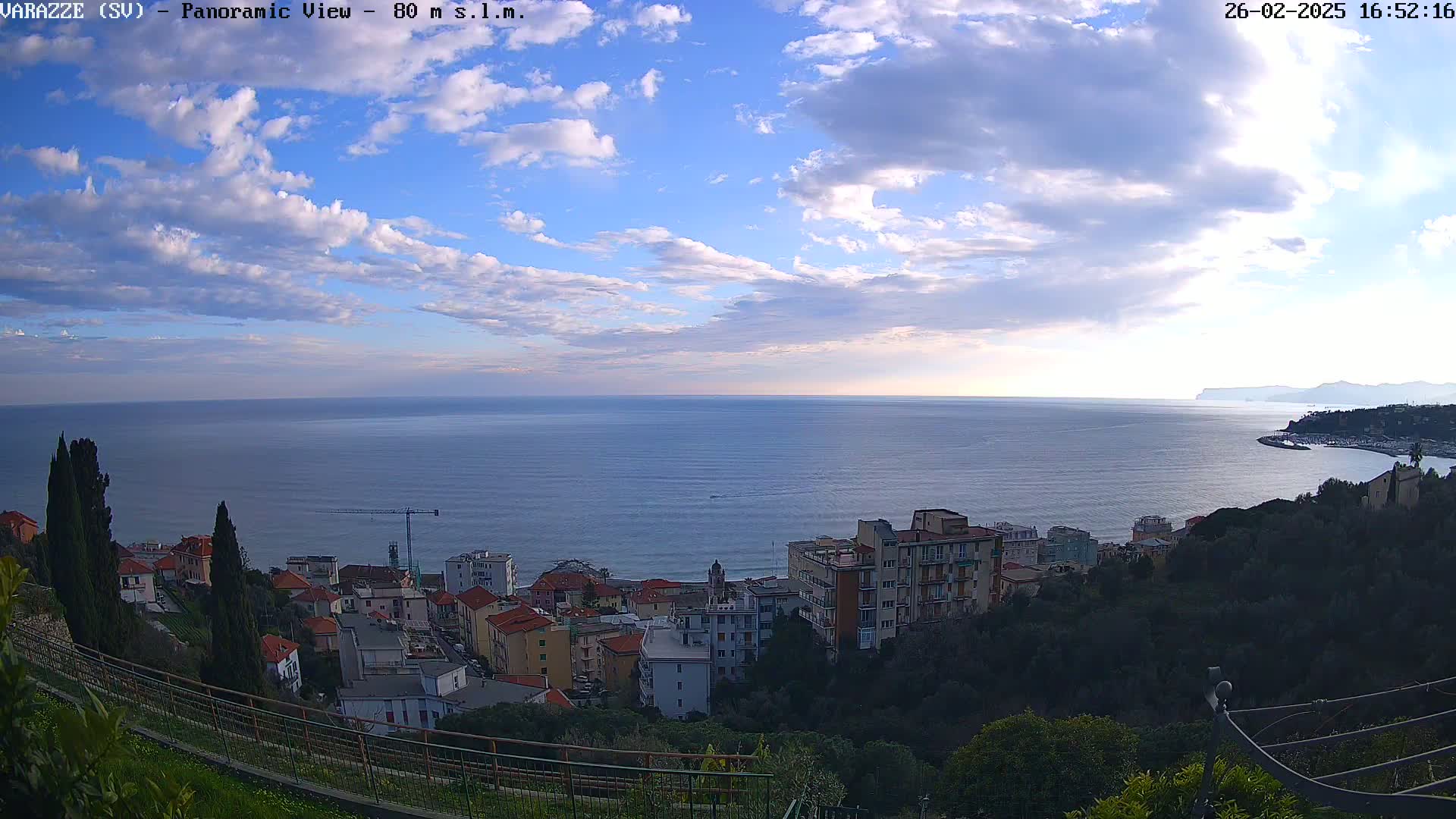 A panoramic view shows a partly cloudy sky over a calm sea, with a coastal town and hills in the foreground.