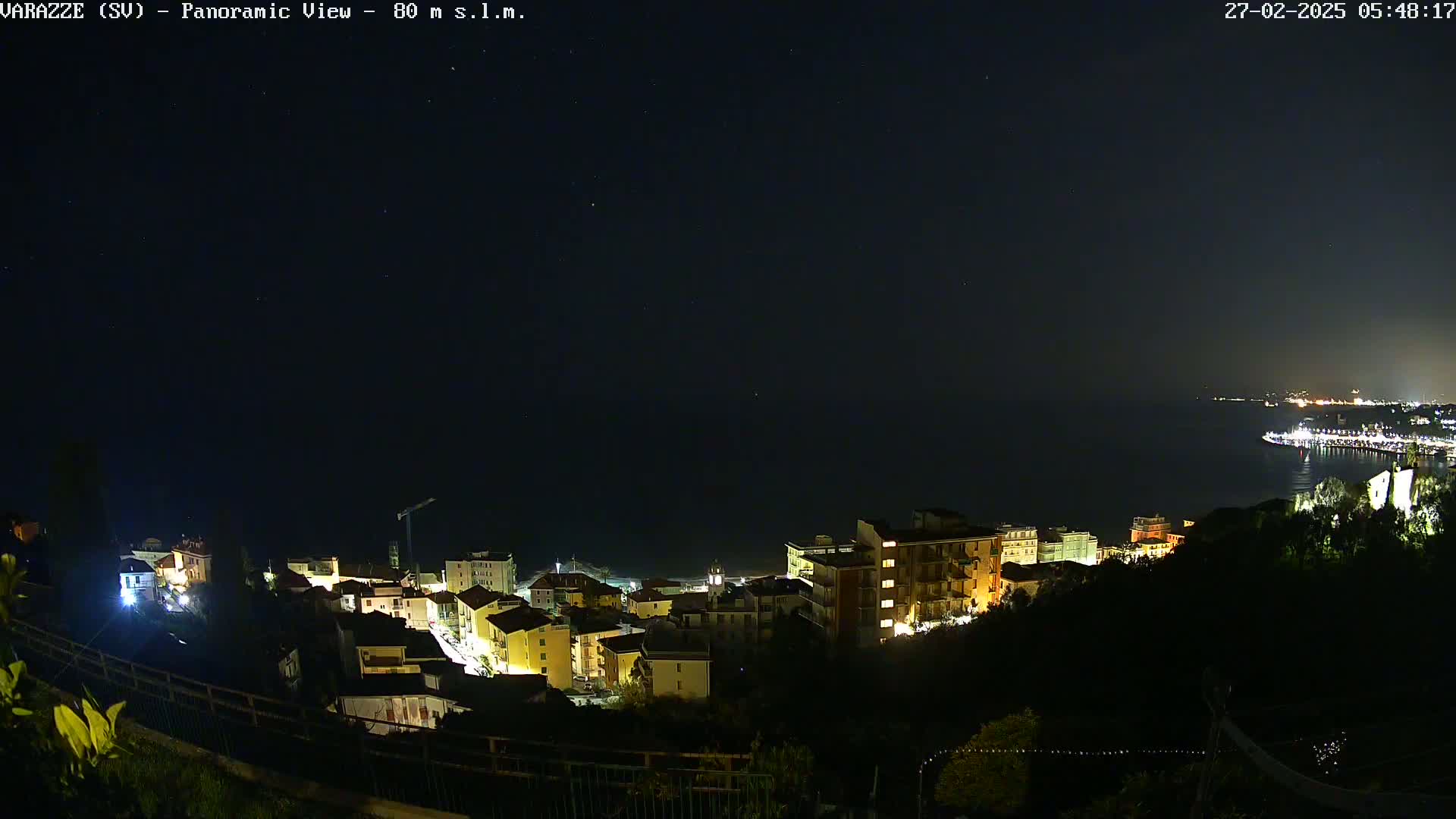 A nighttime, panoramic view of a coastal town with buildings illuminated, overlooking a dark sea under a clear, starry sky.