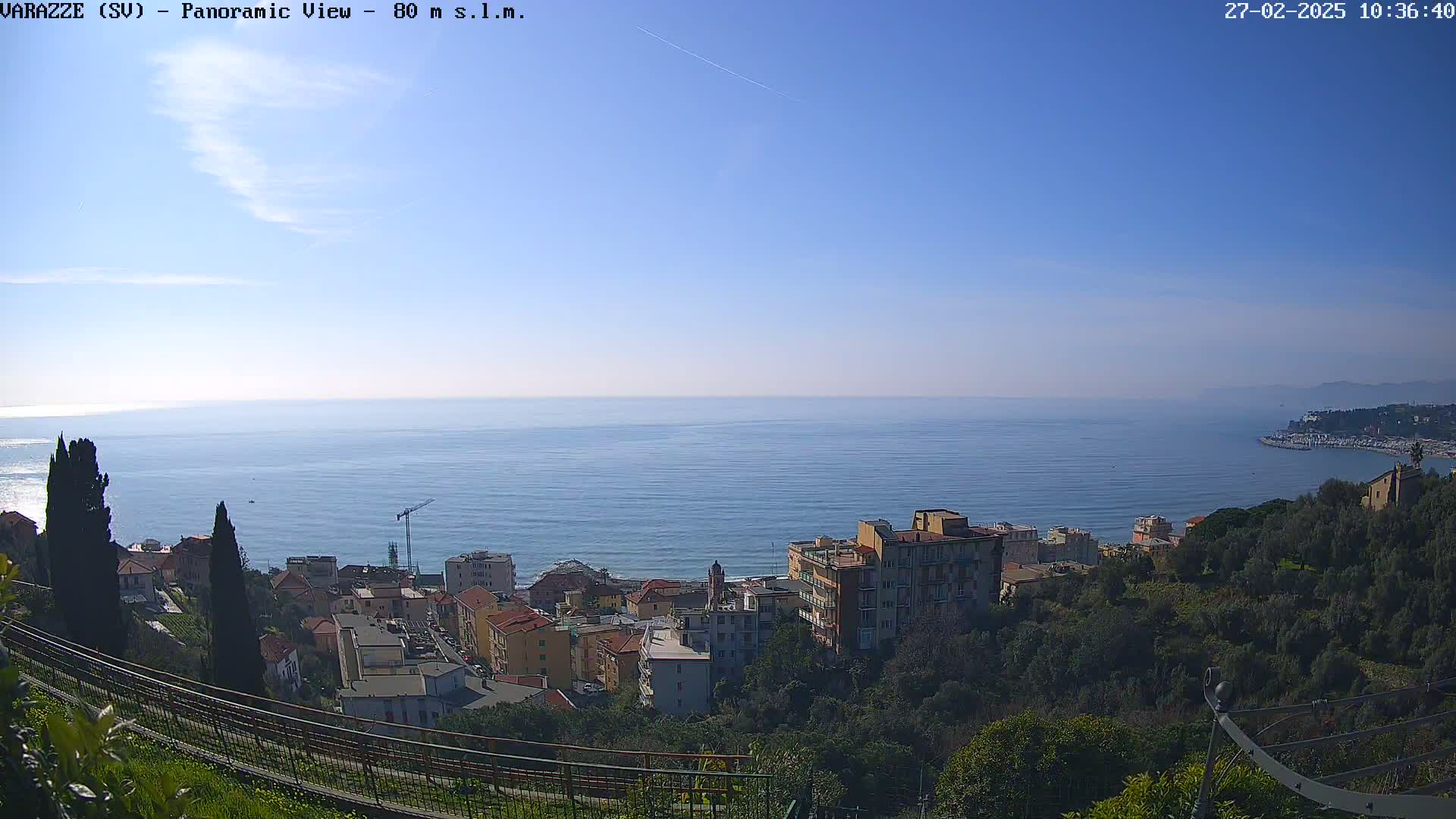 A panoramic view shows a coastal town nestled among green hills overlooking a calm blue sea under a clear blue sky.