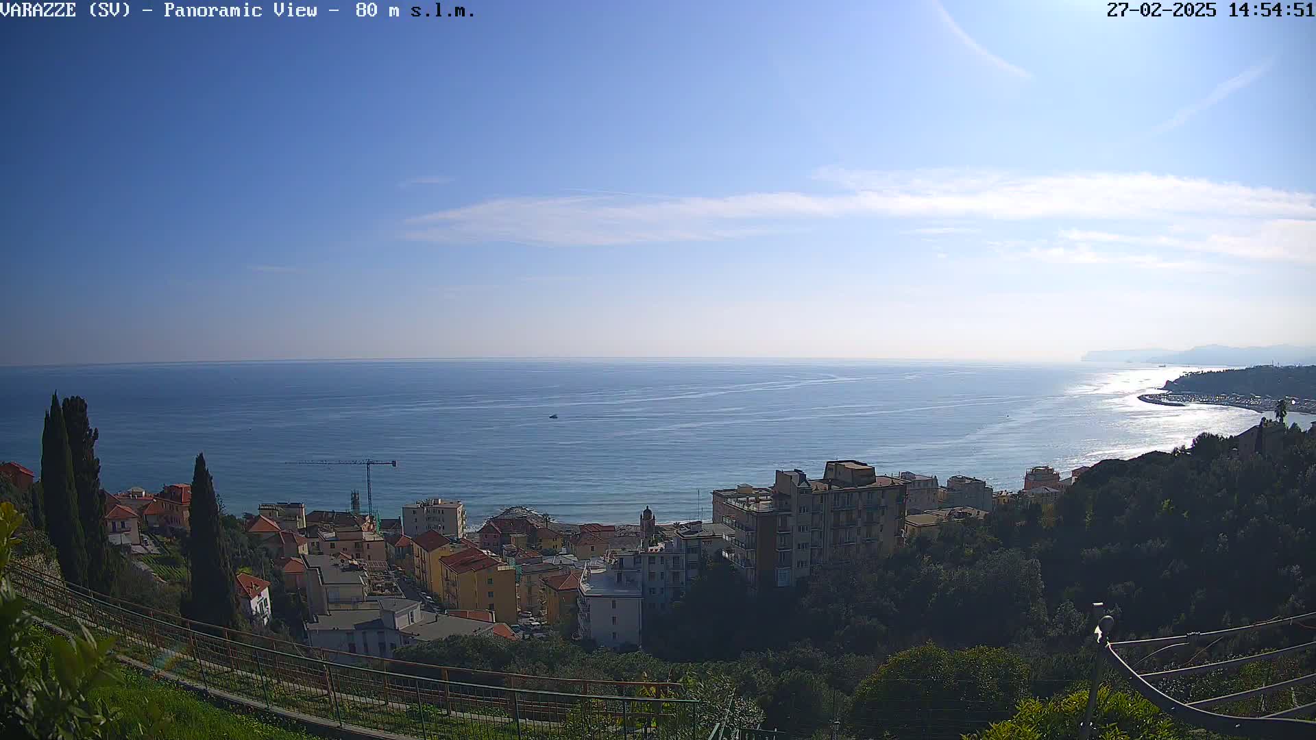 A panoramic view of a coastal town nestled on a hillside overlooking a calm, blue sea under a mostly clear sky.