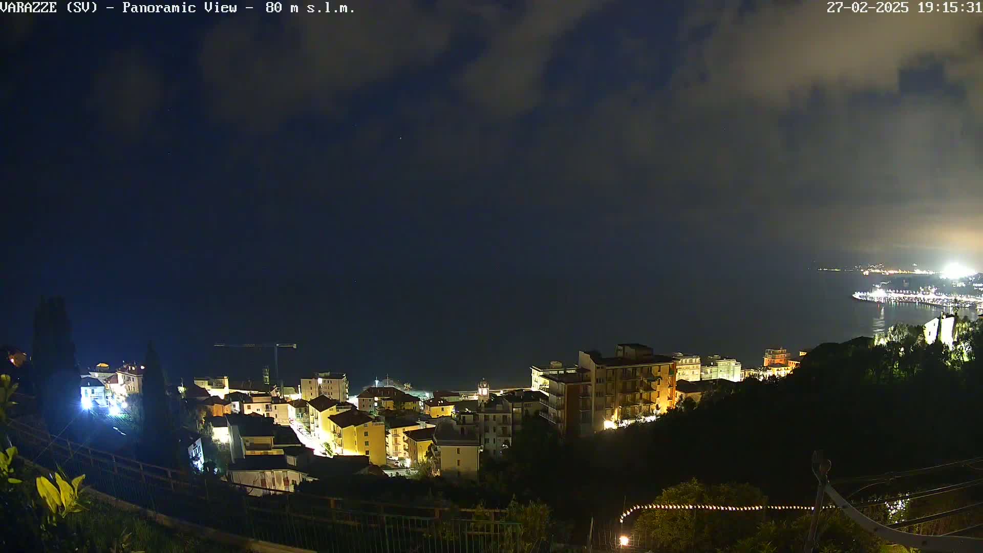 A nighttime panoramic view shows a coastal town with illuminated buildings nestled on a hillside overlooking a dark sea under a cloudy sky.