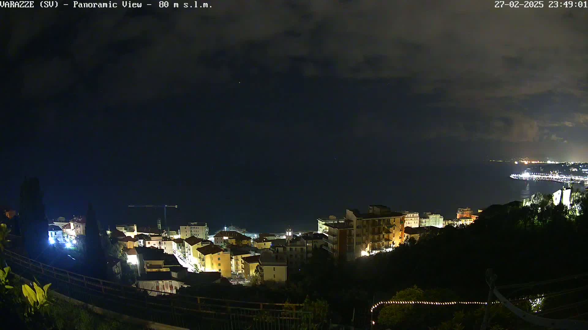 A nighttime panoramic view shows a coastal town illuminated, nestled on a hillside overlooking a dark sea under a partly cloudy sky.