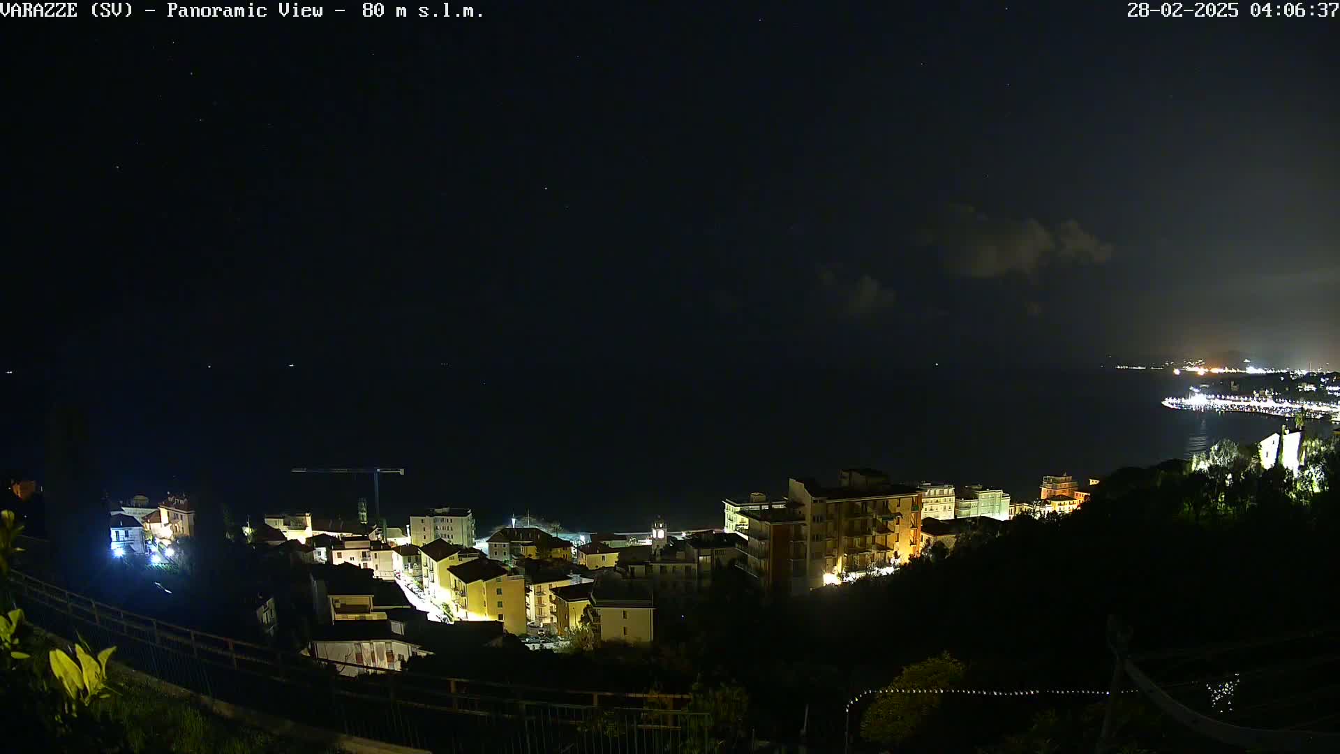 A nighttime panoramic view of a coastal town with lights illuminating buildings, a dark ocean, and a mostly clear sky with a few clouds.
