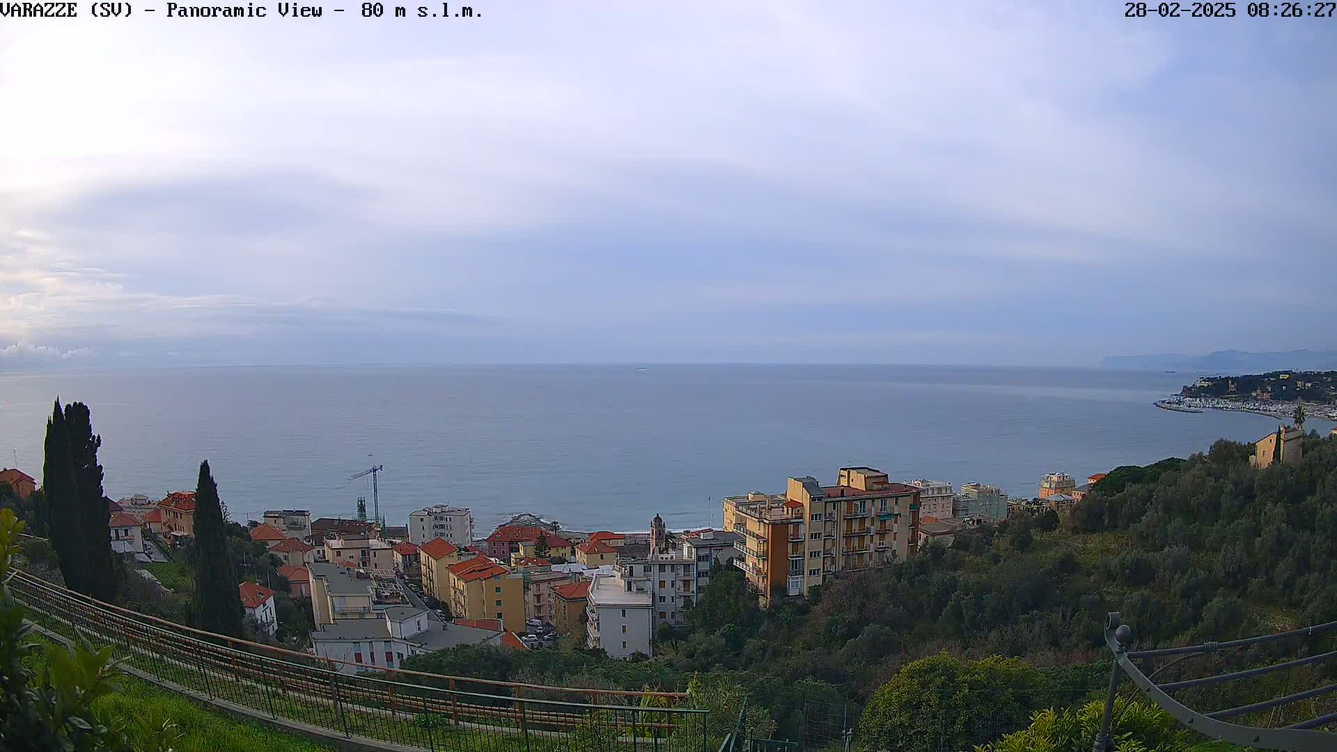 A coastal town nestled on a hillside overlooks a calm sea under a mostly cloudy sky.