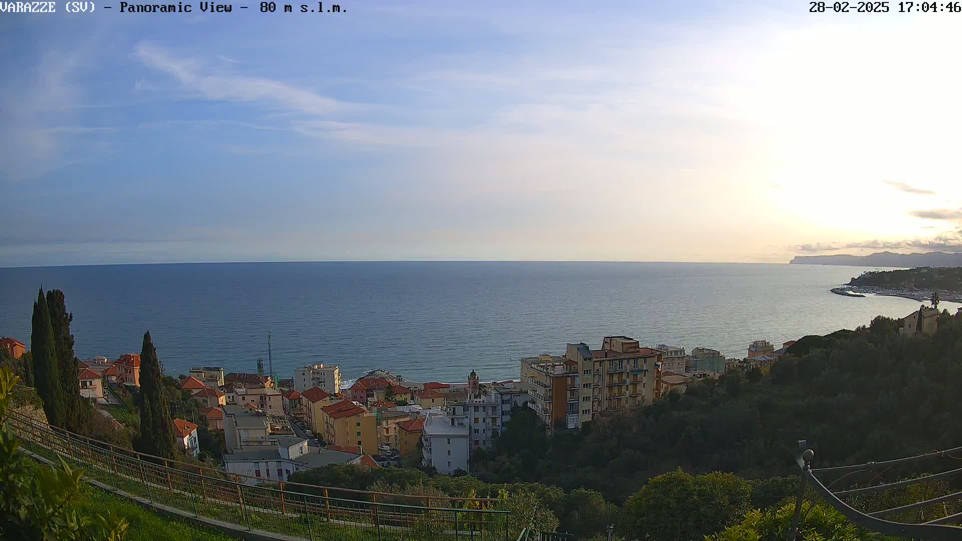 A coastal town with buildings nestled on a hillside overlooking a calm sea under a partly cloudy sky.