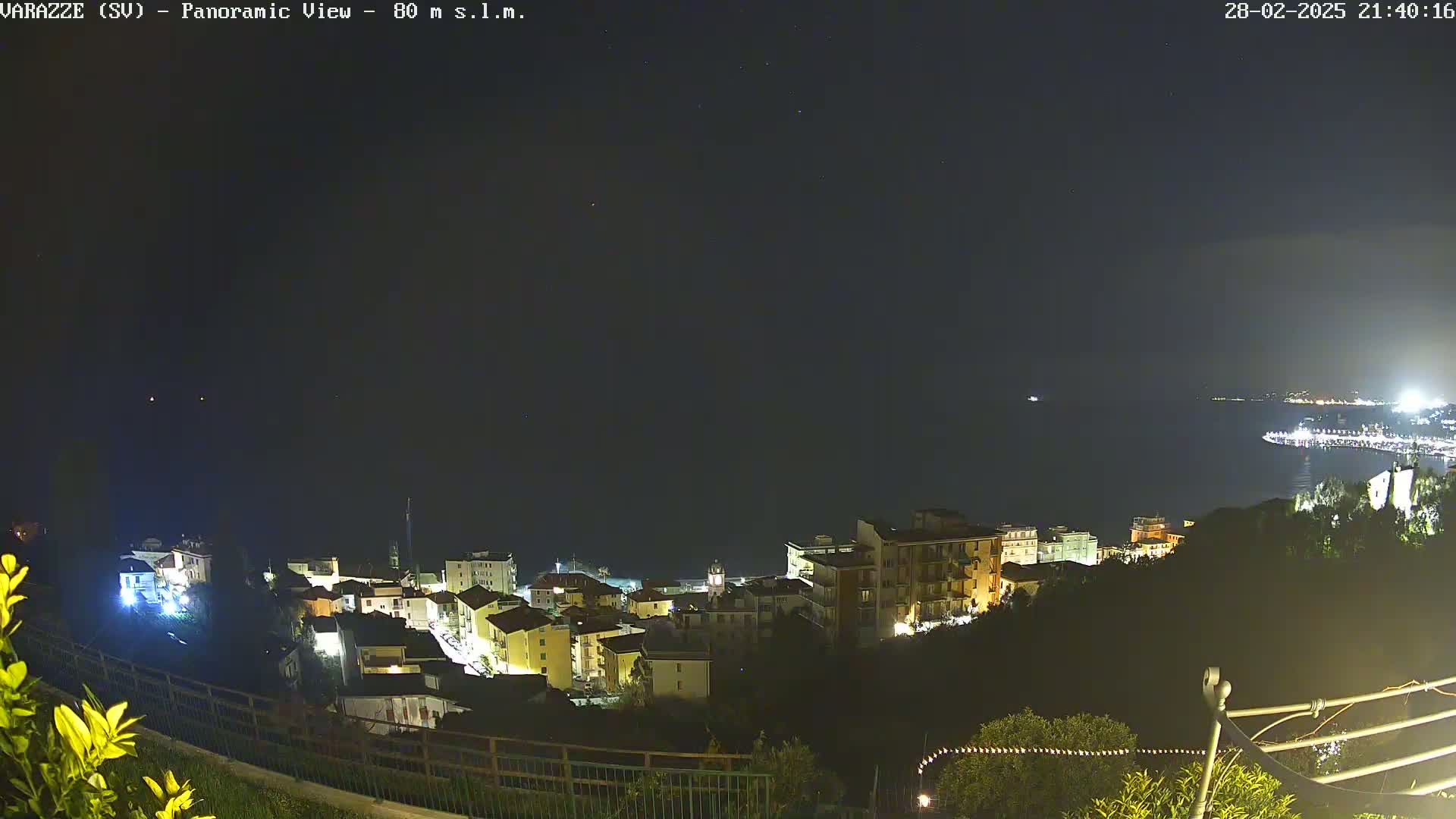 A nighttime, panoramic view of a coastal town with a calm sea, illuminated buildings, and a clear sky speckled with a few stars.