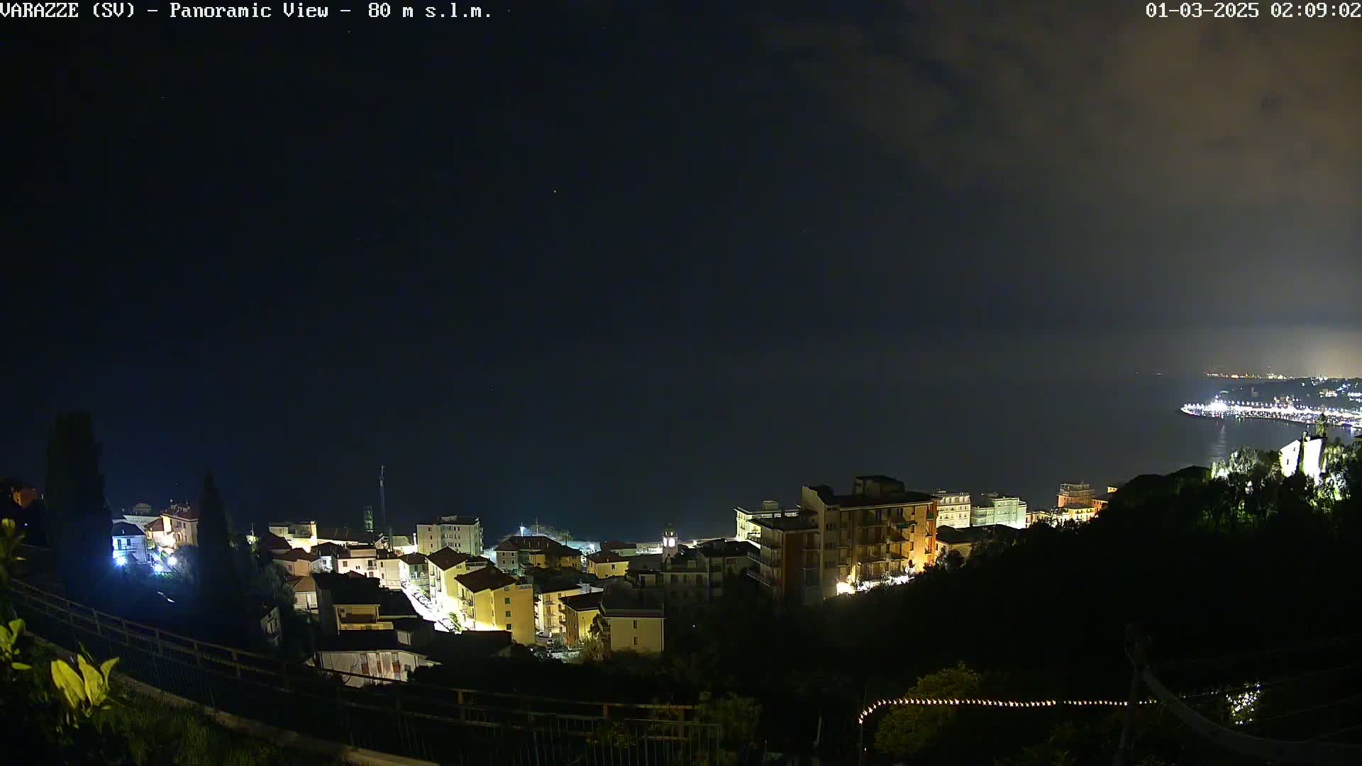 A nighttime, panoramic view of a coastal town with illuminated buildings, overlooking a dark body of water under a mostly clear sky.