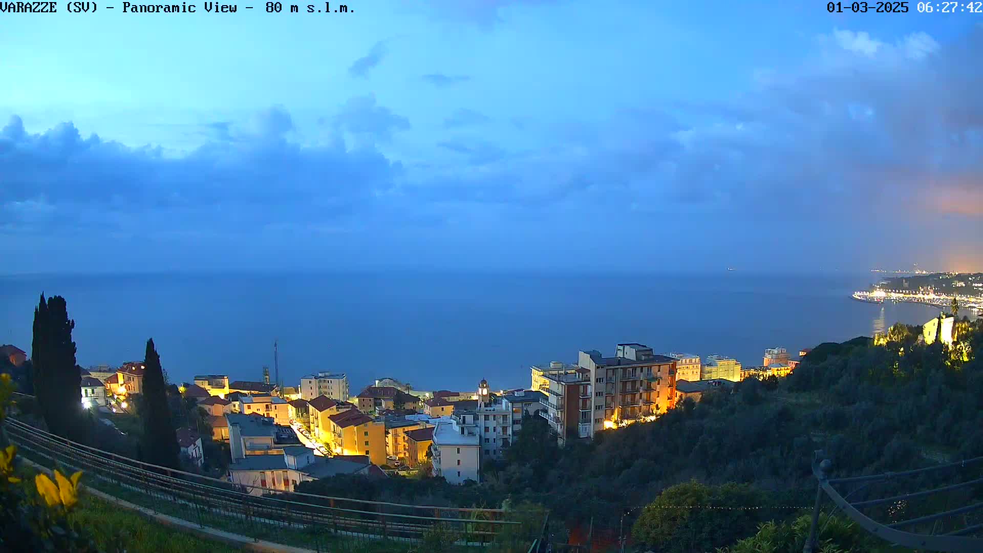 A coastal town at twilight, illuminated with some lights, overlooks a calm sea under a partly cloudy sky.