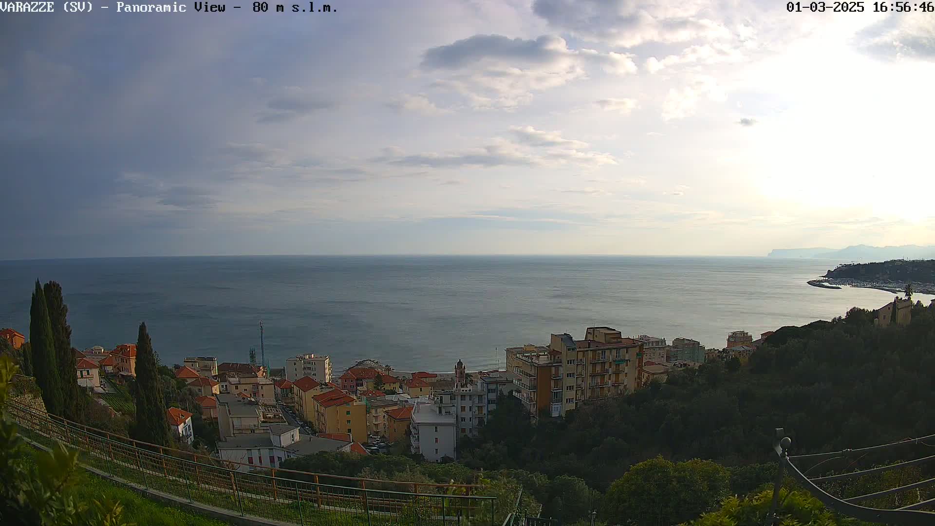 A coastal town with low-rise buildings is nestled in a hillside overlooking a calm sea under a partly cloudy sky.