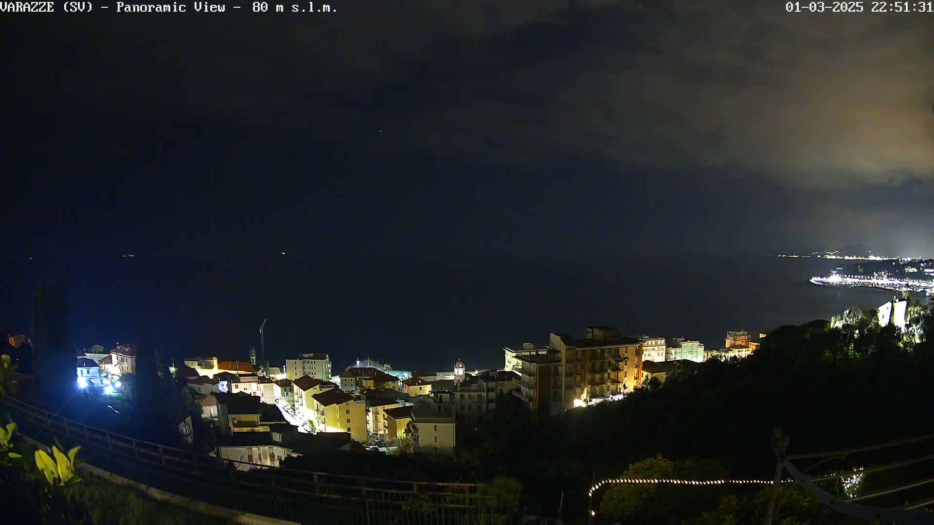 A nighttime panoramic view shows a coastal town illuminated against a dark, cloudy sky and a calm sea.
